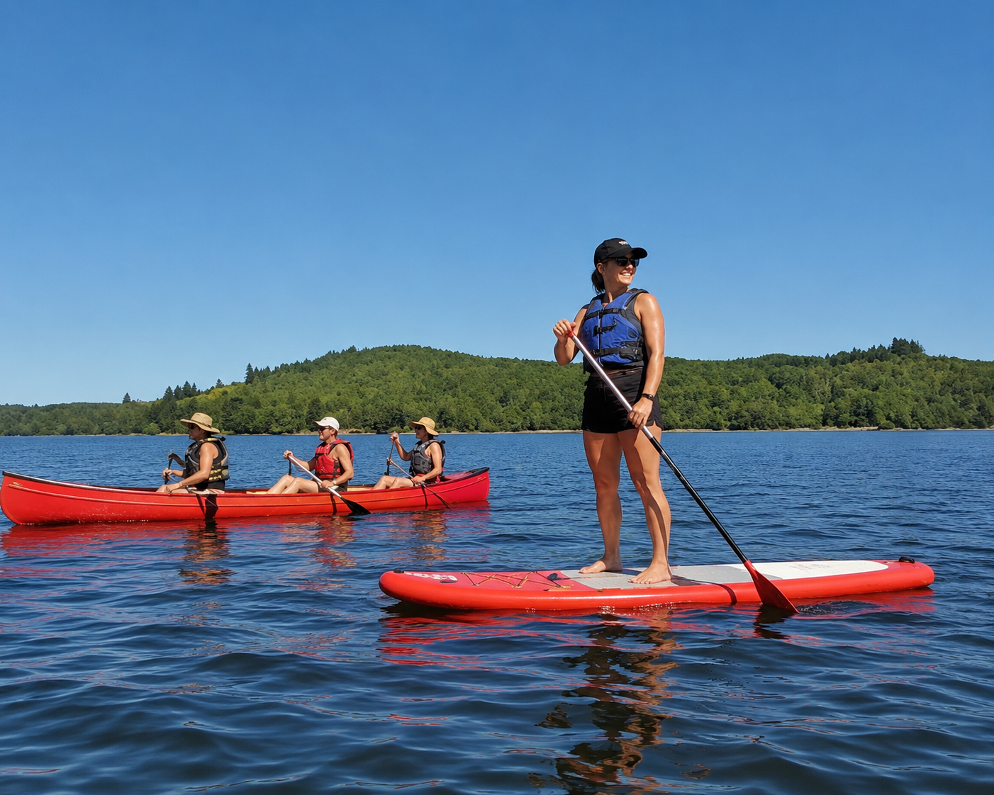 Four people enjoying water activities on a lake, with one person paddleboarding and three people in a kayak, surrounded by green hills and a clear blue sky.