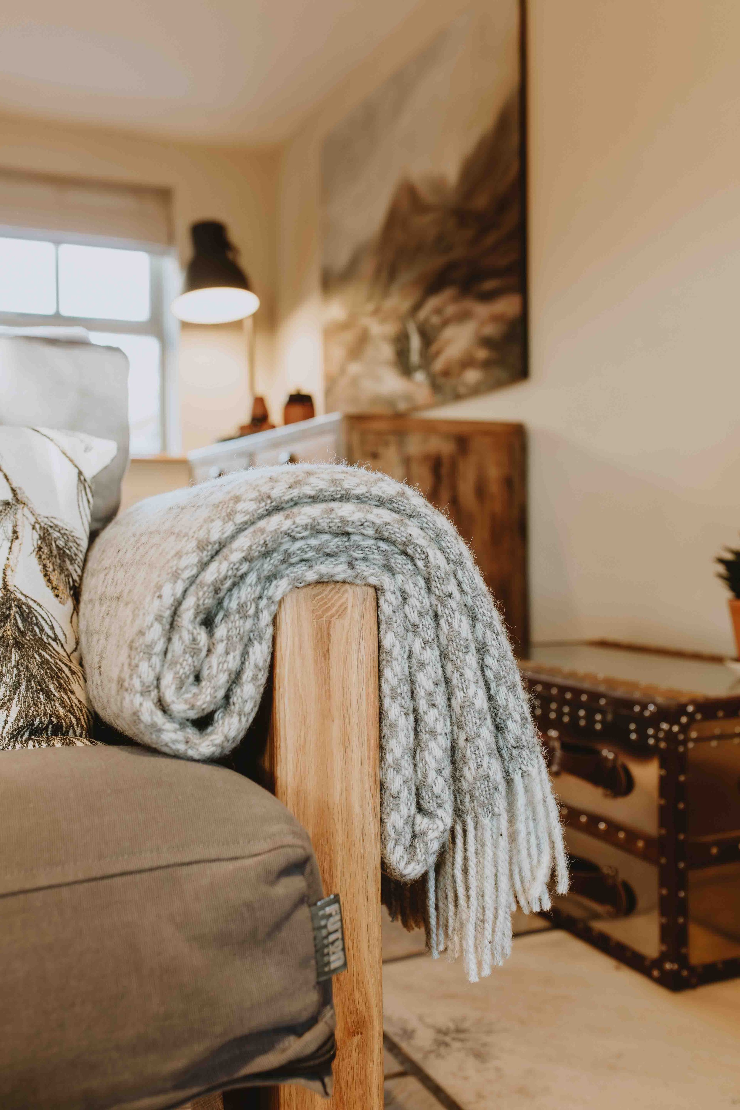 A cozy living room corner with a rolled-up wool blanket draped over a wooden armrest of a sofa. Behind, a table lamp and a framed landscape picture hang on the wall. A window with blinds lets in natural light, and a decorative armchair with nailhead trim is partially visible.