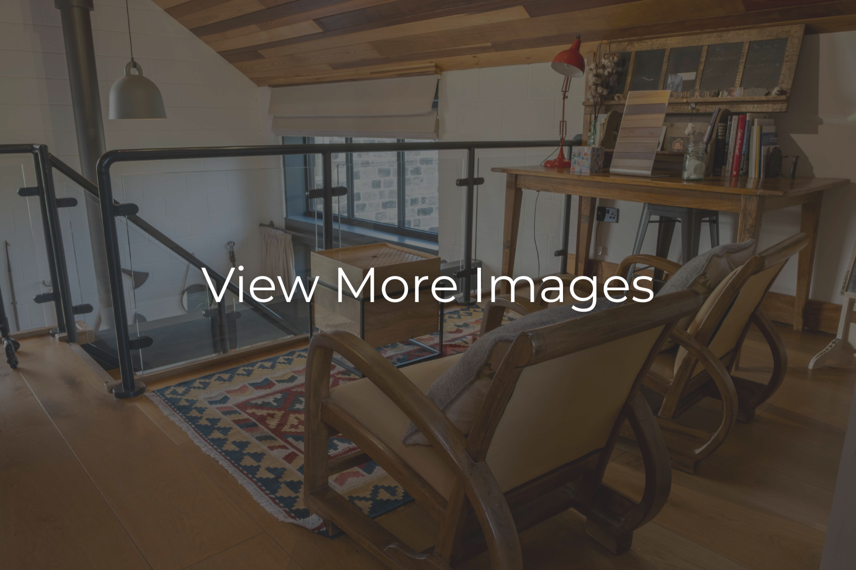 Loft interior with wooden ceiling and flooring, featuring a staircase with glass and metal railing, a wooden desk with books and decor, a vintage rocking chair with cushion, a patterned rug, a window with a shade, and cozy lighting.