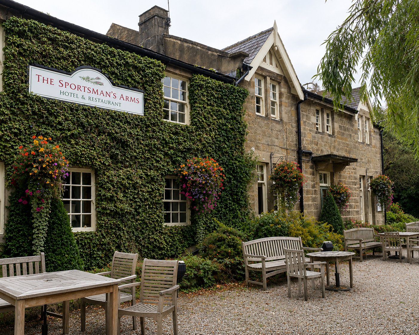 Exterior of The Sportsman's Arms hotel and restaurant with ivy-covered walls, hanging flower baskets, and outdoor seating area with wooden tables and chairs.