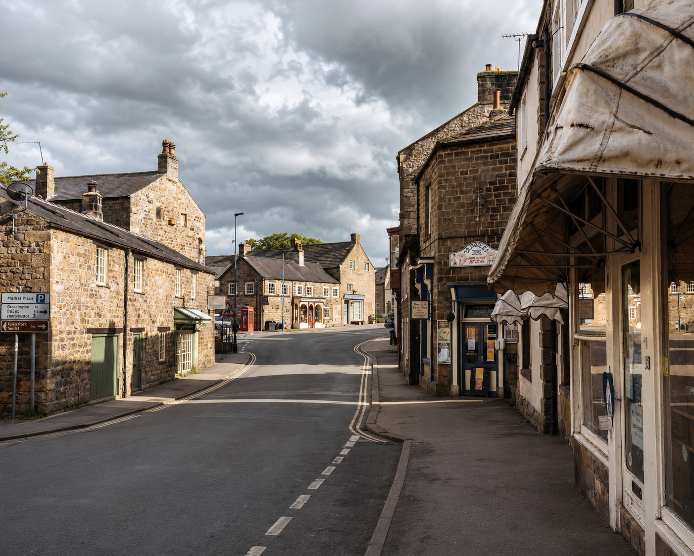 A quiet small town street with stone buildings, a few shops and a street sign, under cloudy skies.