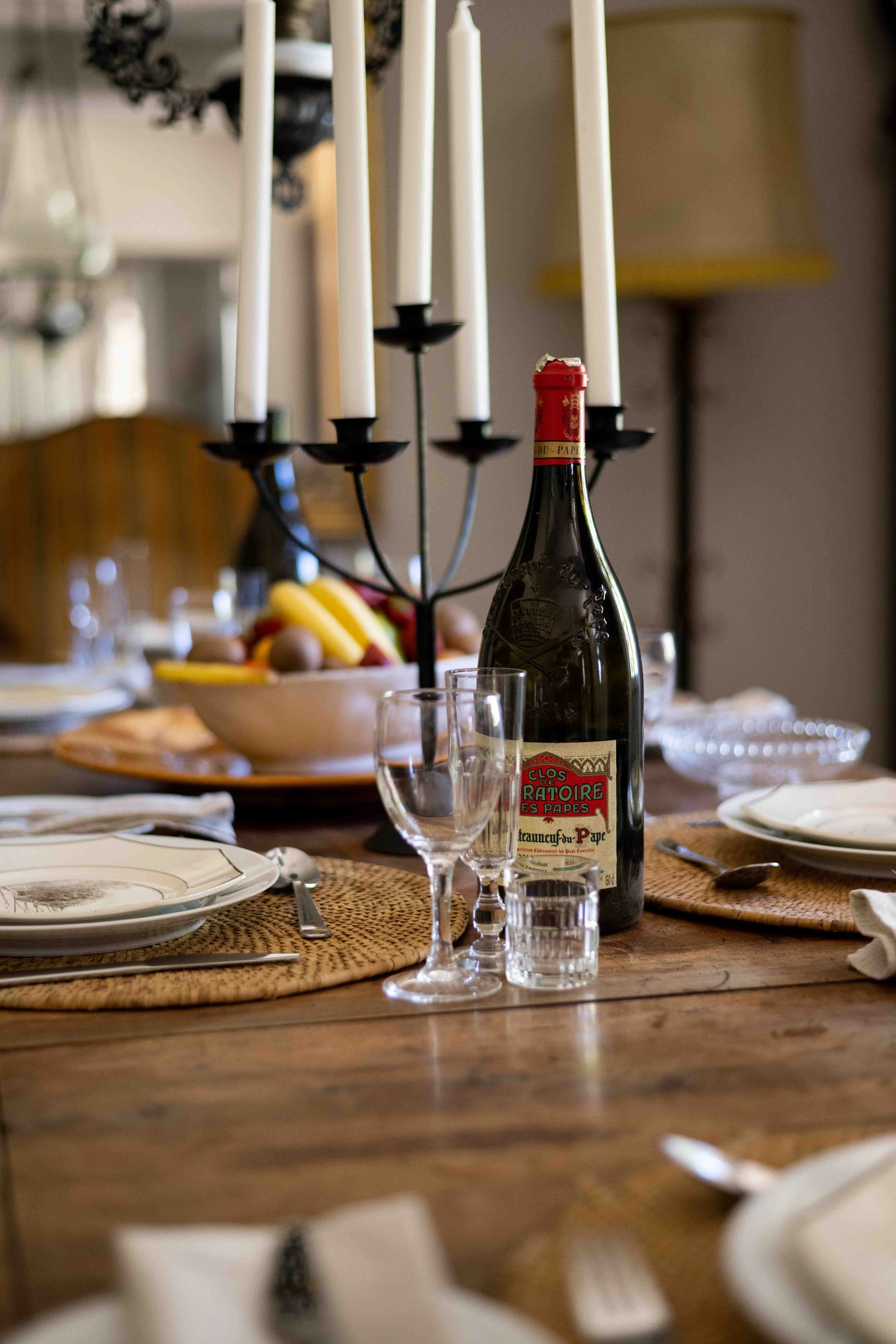 A dining table set with plates, glasses, cutlery, and woven placemats, with a bottle of red wine and a candelabra with unlit white candles. A fruit bowl and a table lamp are also visible in the background.