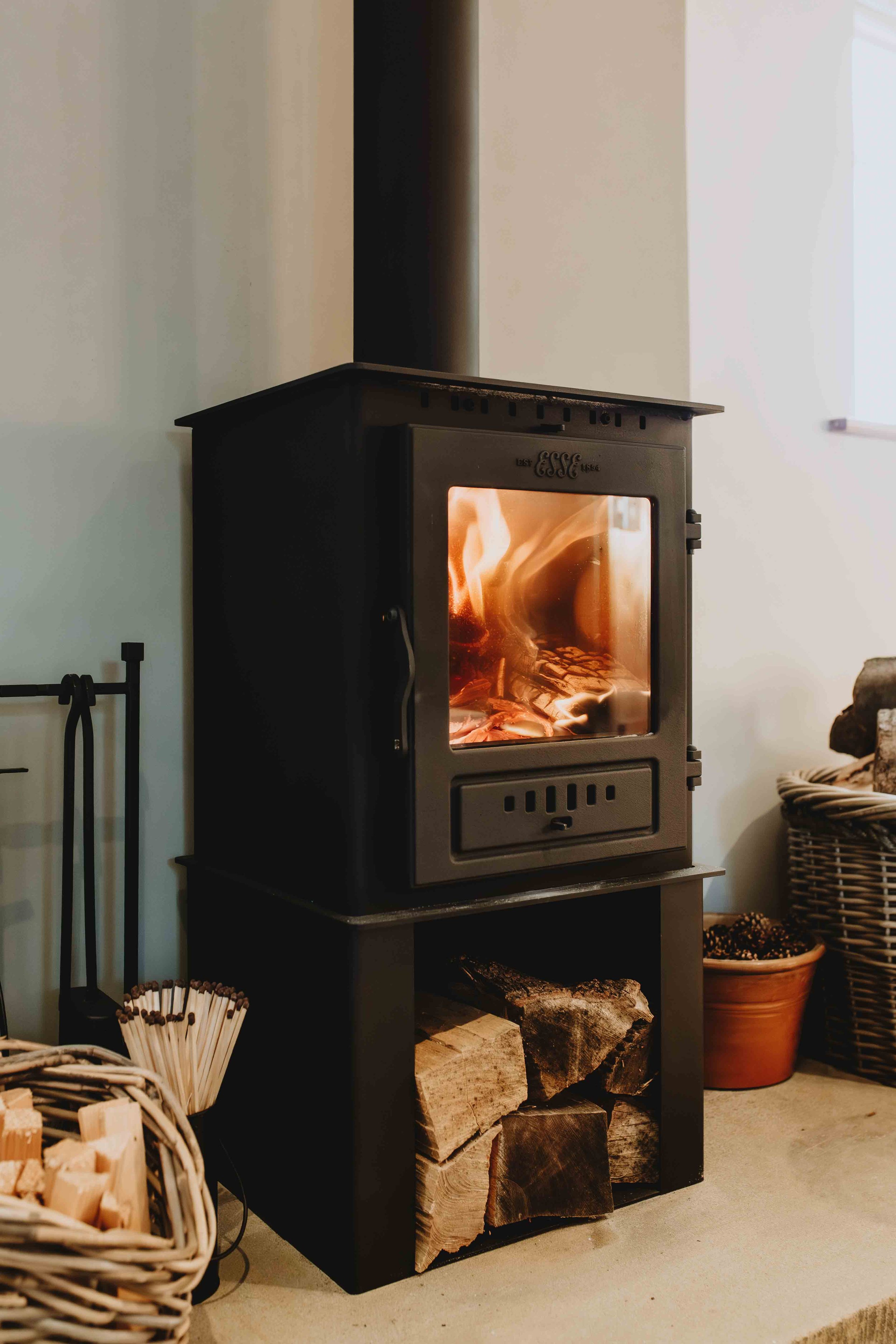 A black wood-burning stove with a glass door showing flames inside, set against a white wall with a window on the right. Firewood is stored inside and below the stove, and there are baskets and fireplace tools nearby.