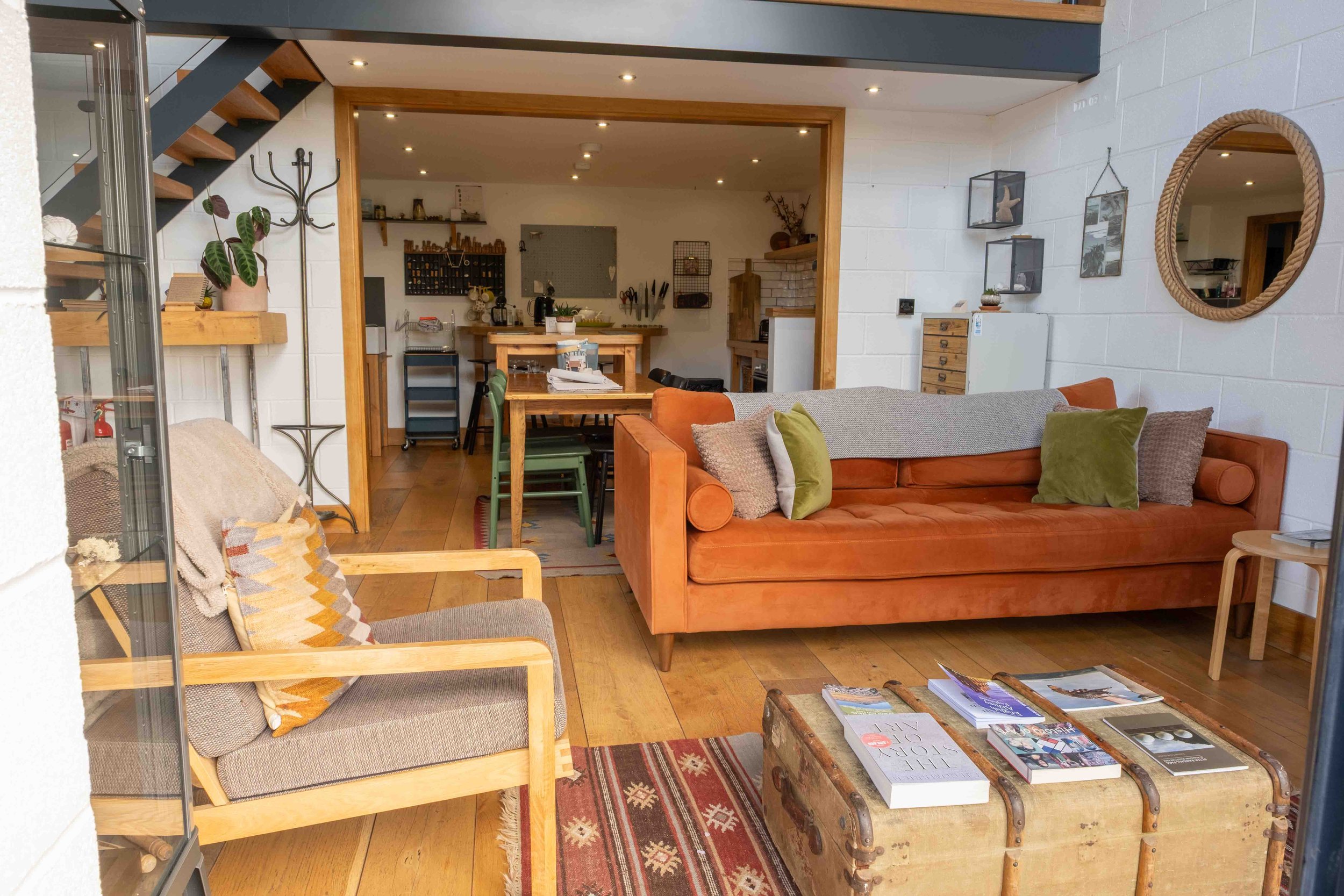 Living room with a sofa and a wooden coffee table displaying magazines. Behind the sofa is a white brick wall with round mirrors and wall shelves. The adjacent kitchen has a dining table with chairs, a kitchen counter, and various appliances, with a staircase leading upstairs.