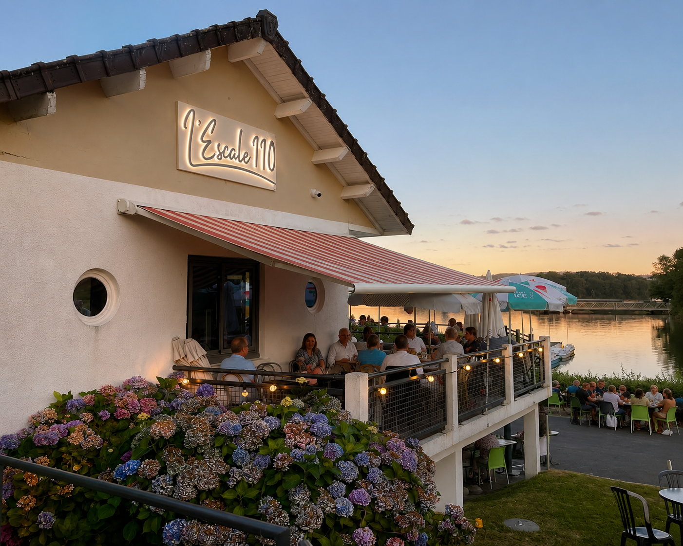 The image shows a two-story restaurant building with a sign that reads 'Escale 10,' nestled beside a river during sunset. The outdoor dining patio is filled with people enjoying a meal, and the patio is decorated with string lights and umbrellas. In front of the building, there's a cluster of colorful hydrangea flowers, and in the background, the calm river reflects the sunset sky.
