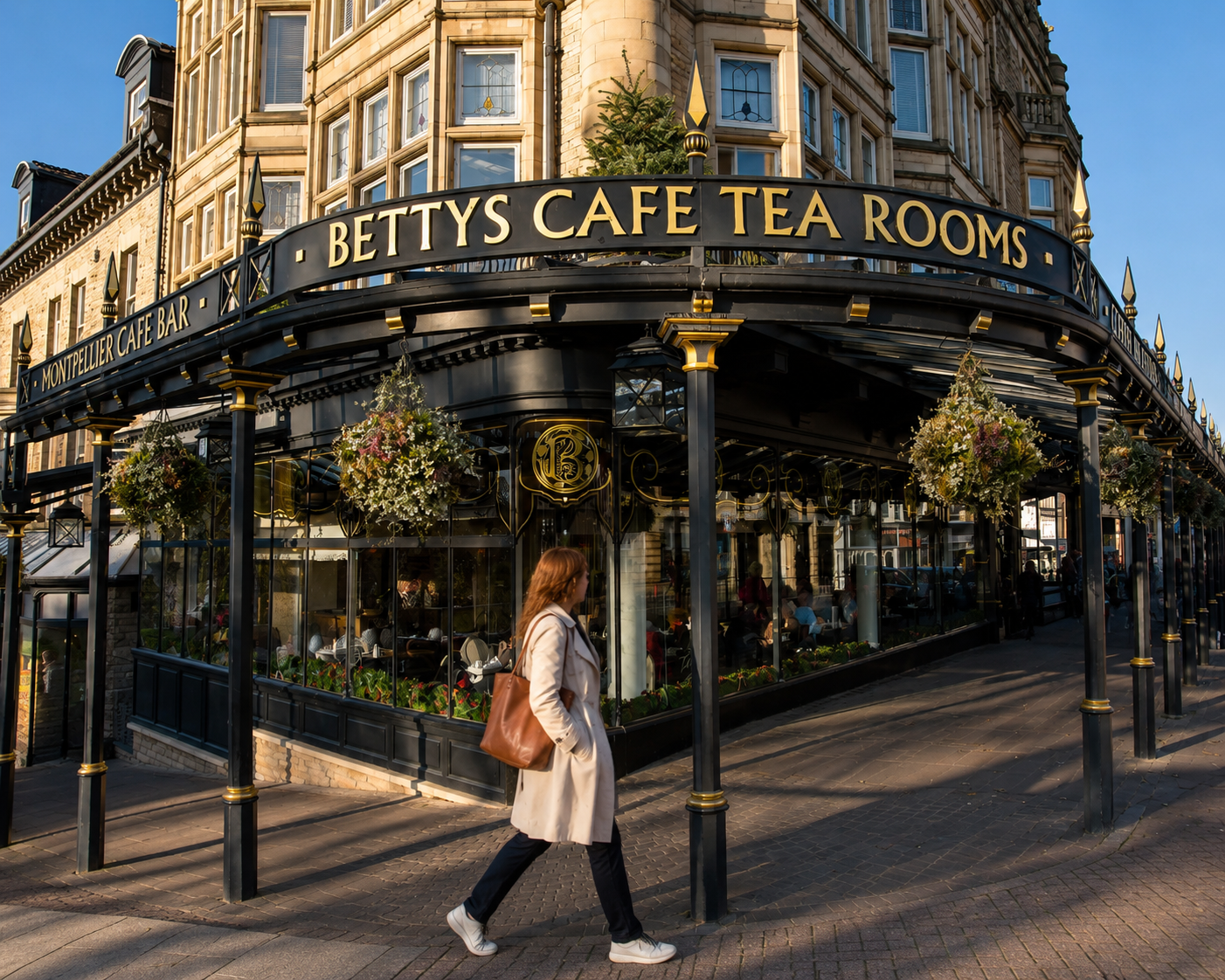 A woman with red hair, wearing a beige coat and white sneakers, walking past a corner café with black and gold signage reading 'Betty's Cafe Tea Rooms'. The café has large glass windows with hanging flower baskets outside, and a building with ornate architecture and multiple windows in the background.