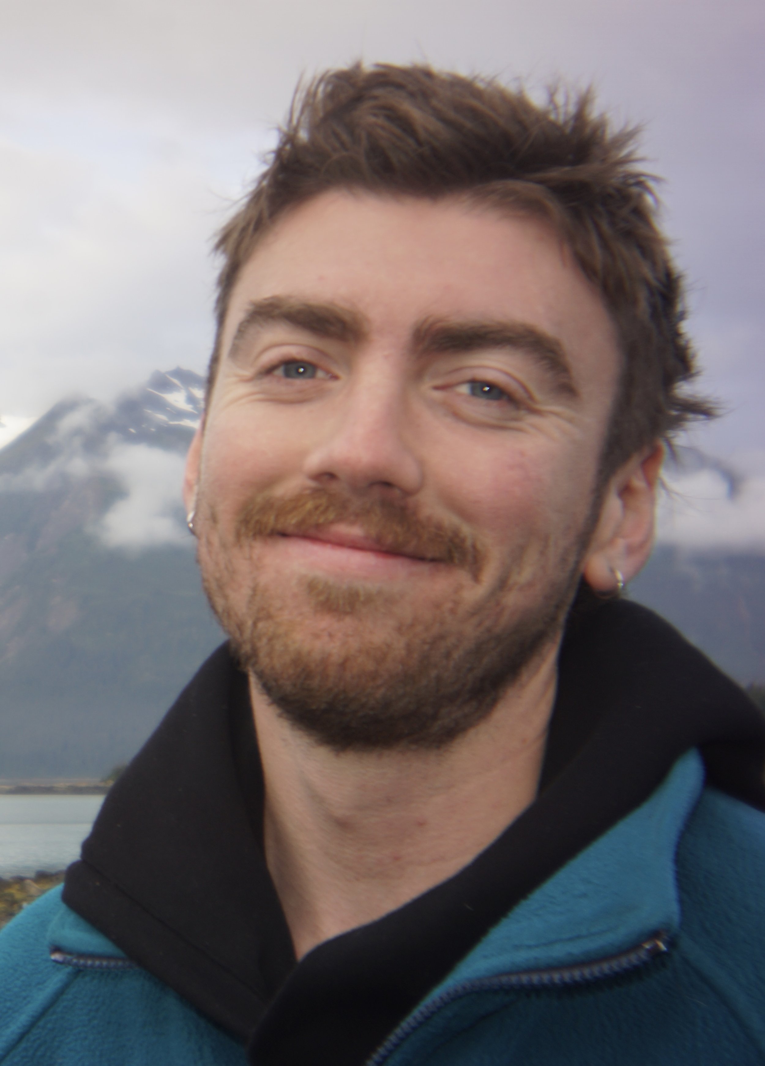A smiling man with brown hair, blue eyes, and a beard in an outdoor setting with mountains and water in the background.