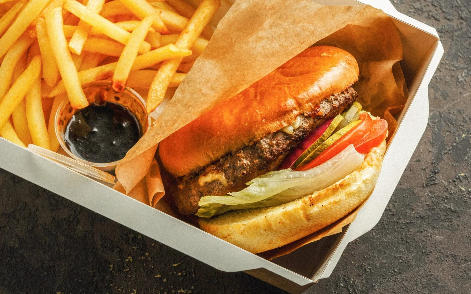 A meal in a takeout box including a burger with lettuce, tomato, pickles, and melted cheese, a serving of French fries, and a small container of ketchup.