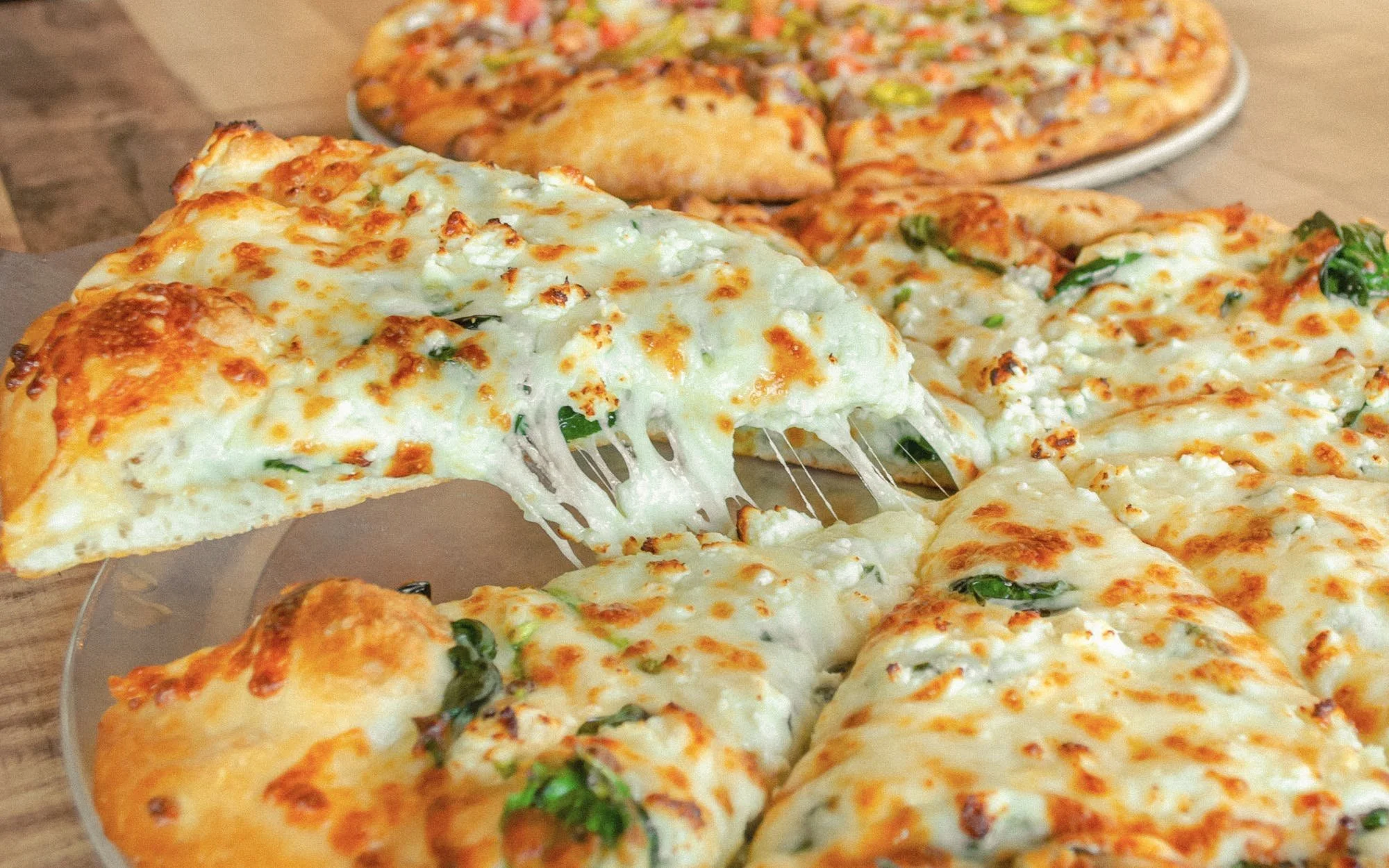 Close-up image of a cheese pizza with a slice being pulled away, revealing melted cheese stretching. In the background, there is a whole pizza topped with various ingredients and another partially visible pizza. The pizzas are on a wooden surface.