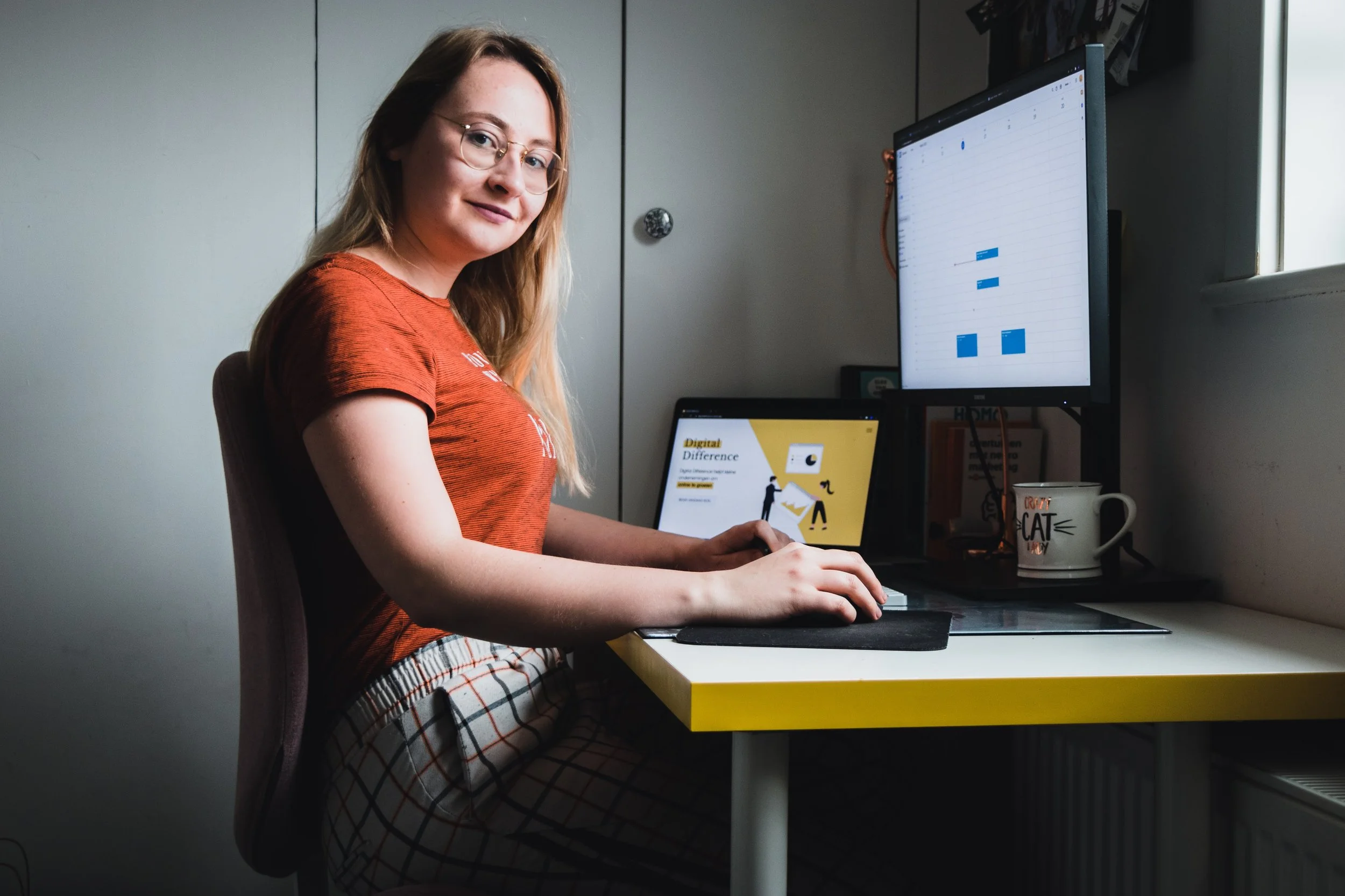 A woman with glasses and long hair working on a computer at a desk in a small office or home workspace. She is wearing a rust-colored shirt and plaid pants, with a smartphone and a calendar or planner on the desk. There are two monitors, one showing a spreadsheet and the other with a presentation slide titled 'Digital Difference'.