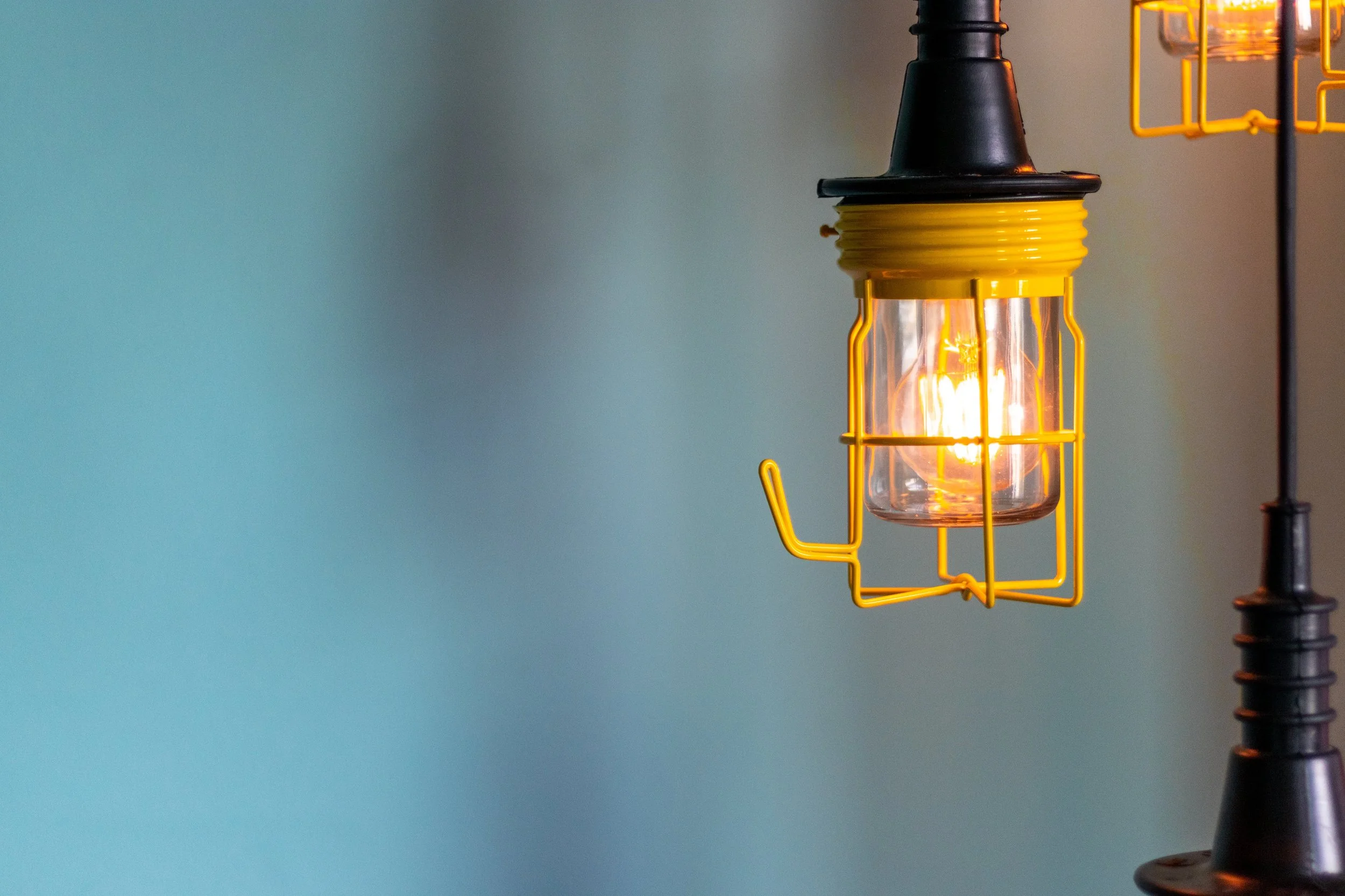 Close-up of a hanging yellow industrial-style lamp with an exposed filament bulb against a plain blue background.