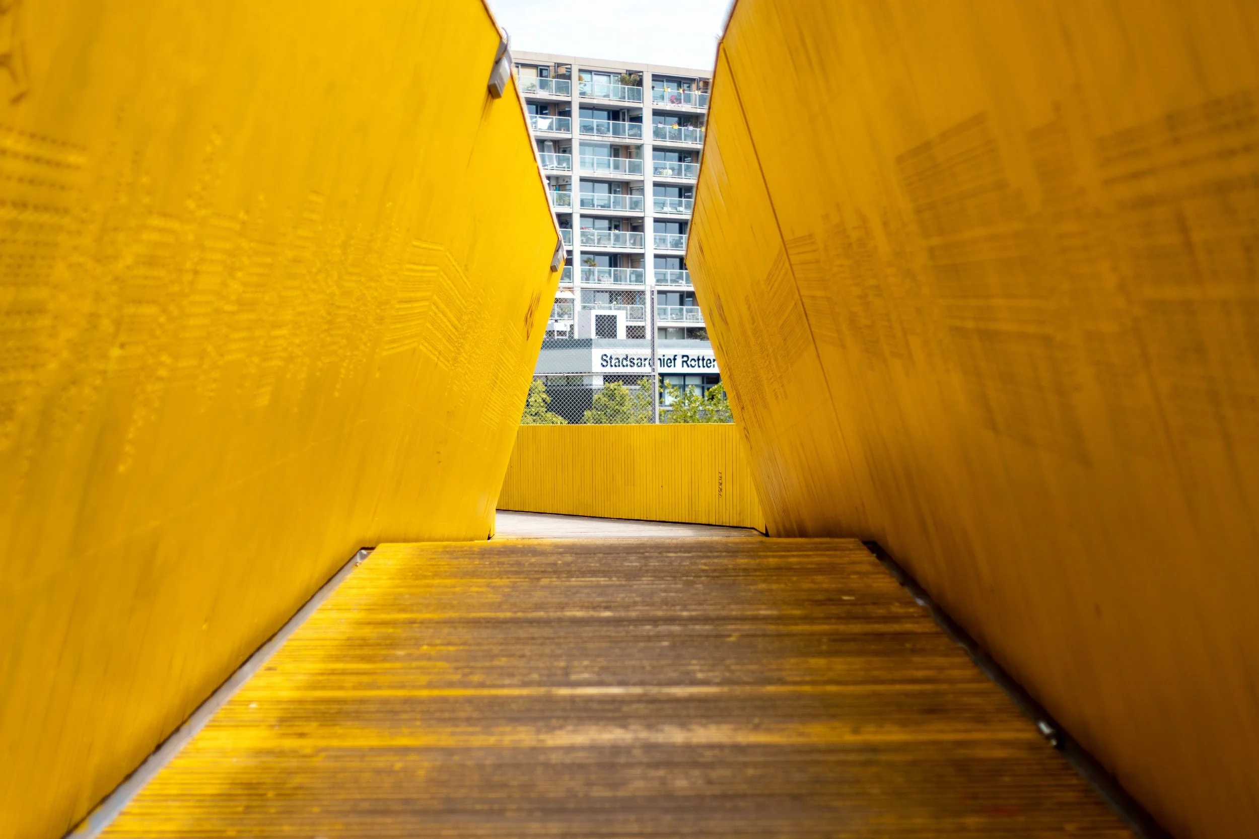 A view from between two yellow wooden structures, looking toward a modern multi-story apartment building with balconies.