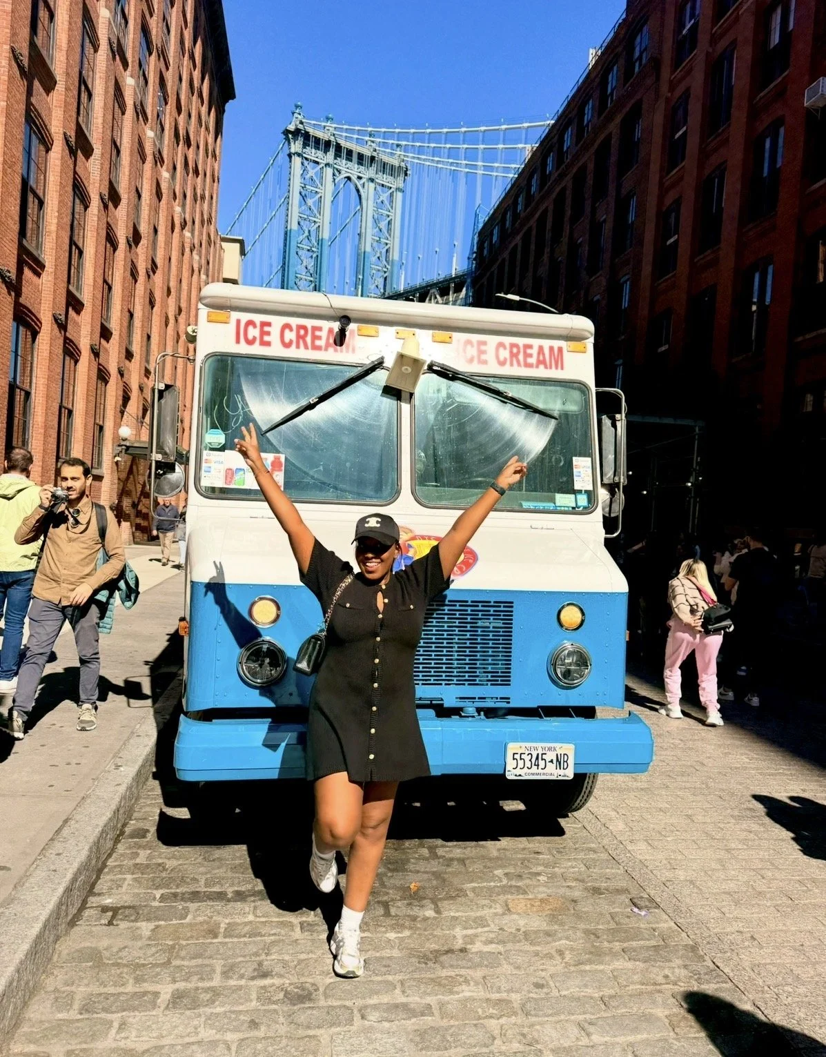 A woman in a black dress and white sneakers celebrating in front of a blue and white ice cream truck on a city street, with the Brooklyn Bridge visible in the background.