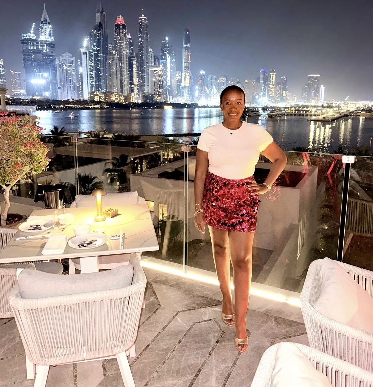 A woman standing on a balcony at night with a city skyline and water in the background, dining table with plates and glasses is in the foreground.