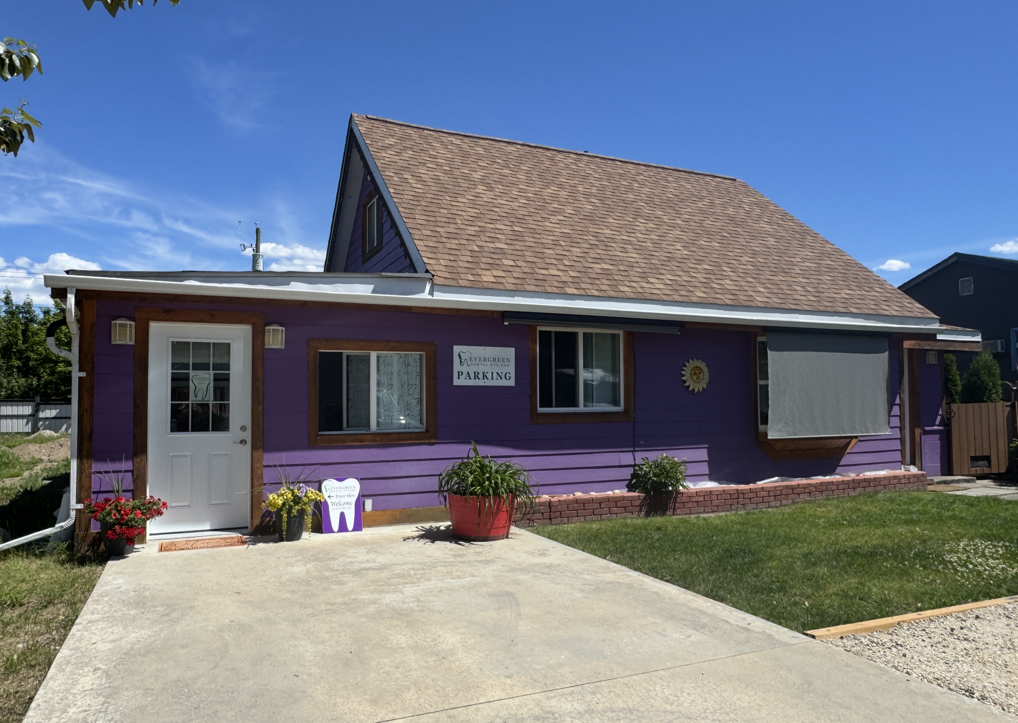 A purple house with a steep gable roof, white door, and large front window. There are potted plants and flowers outside, and a sign indicating parking. The sky is clear and blue.