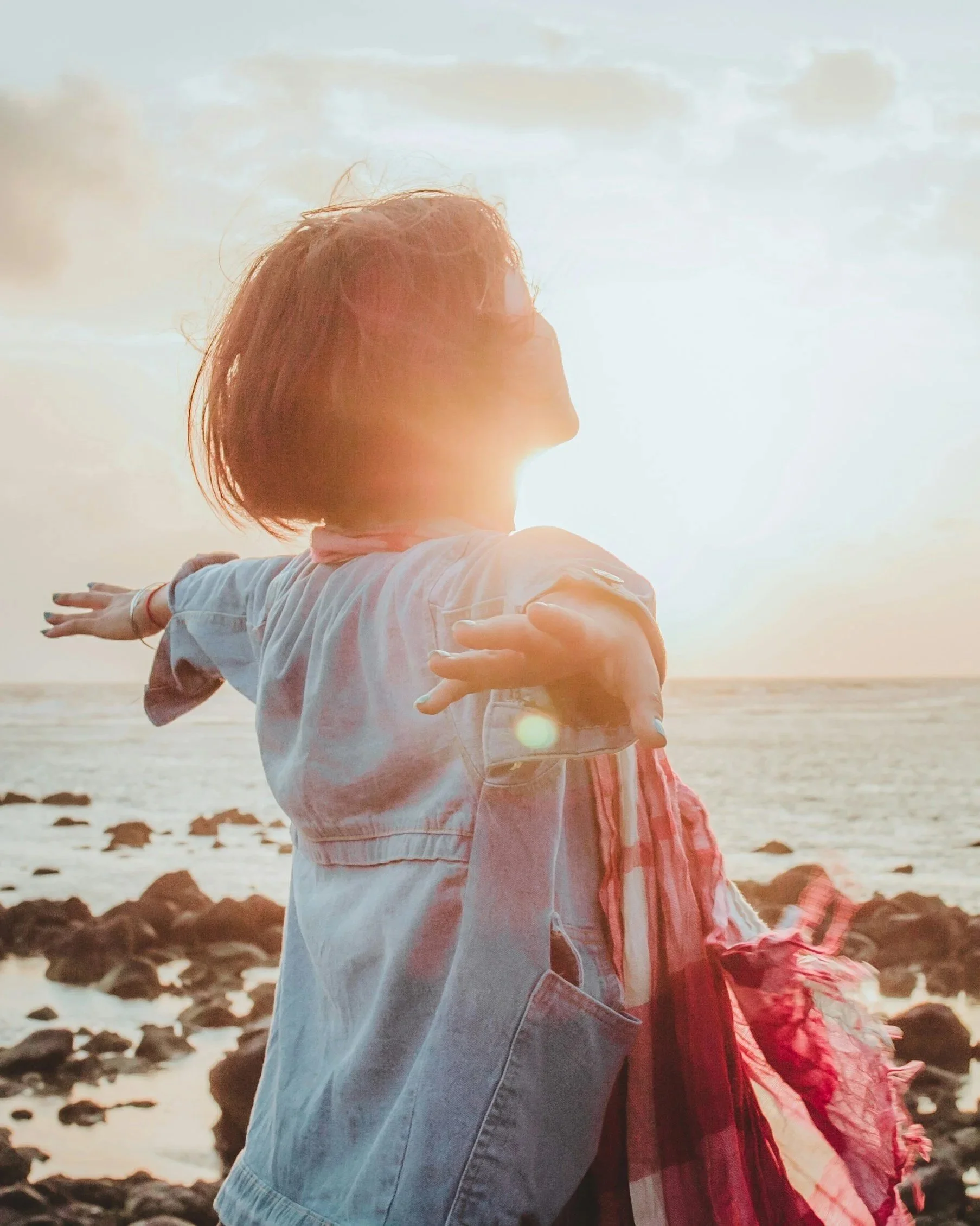 A woman with short hair is standing on a rocky beach at sunset, extending her arms outward as a child with long hair in a plaid shirt is close behind her. The bright sunlight is behind her, creating a warm glow.