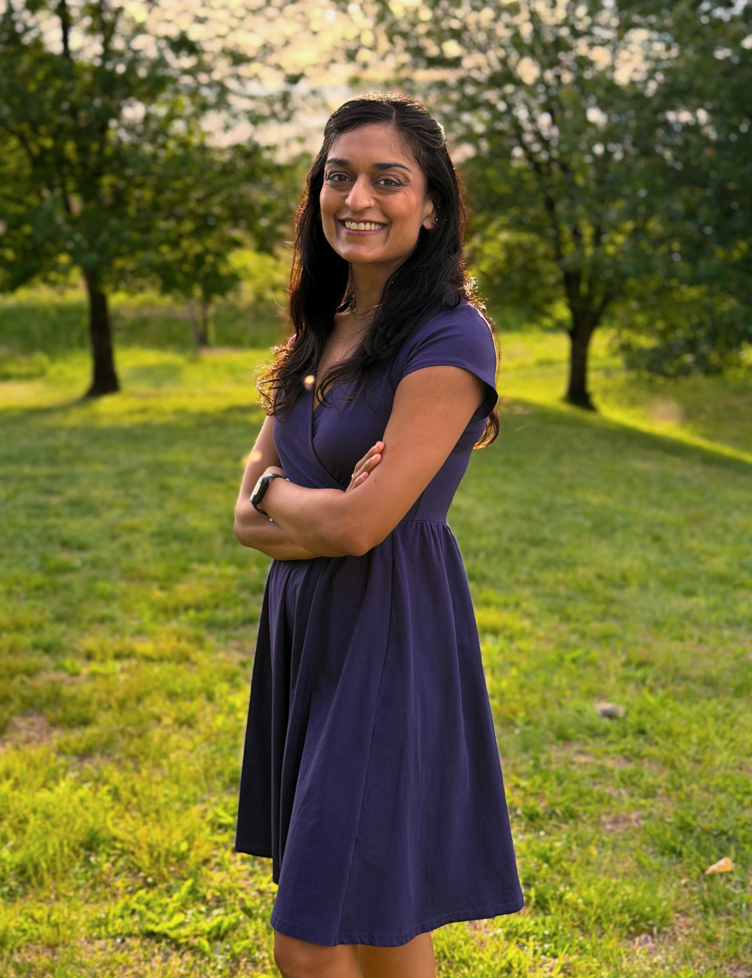A woman with long dark hair, wearing a purple dress, standing outdoors in a park with trees and green grass, smiling and crossing her arms.