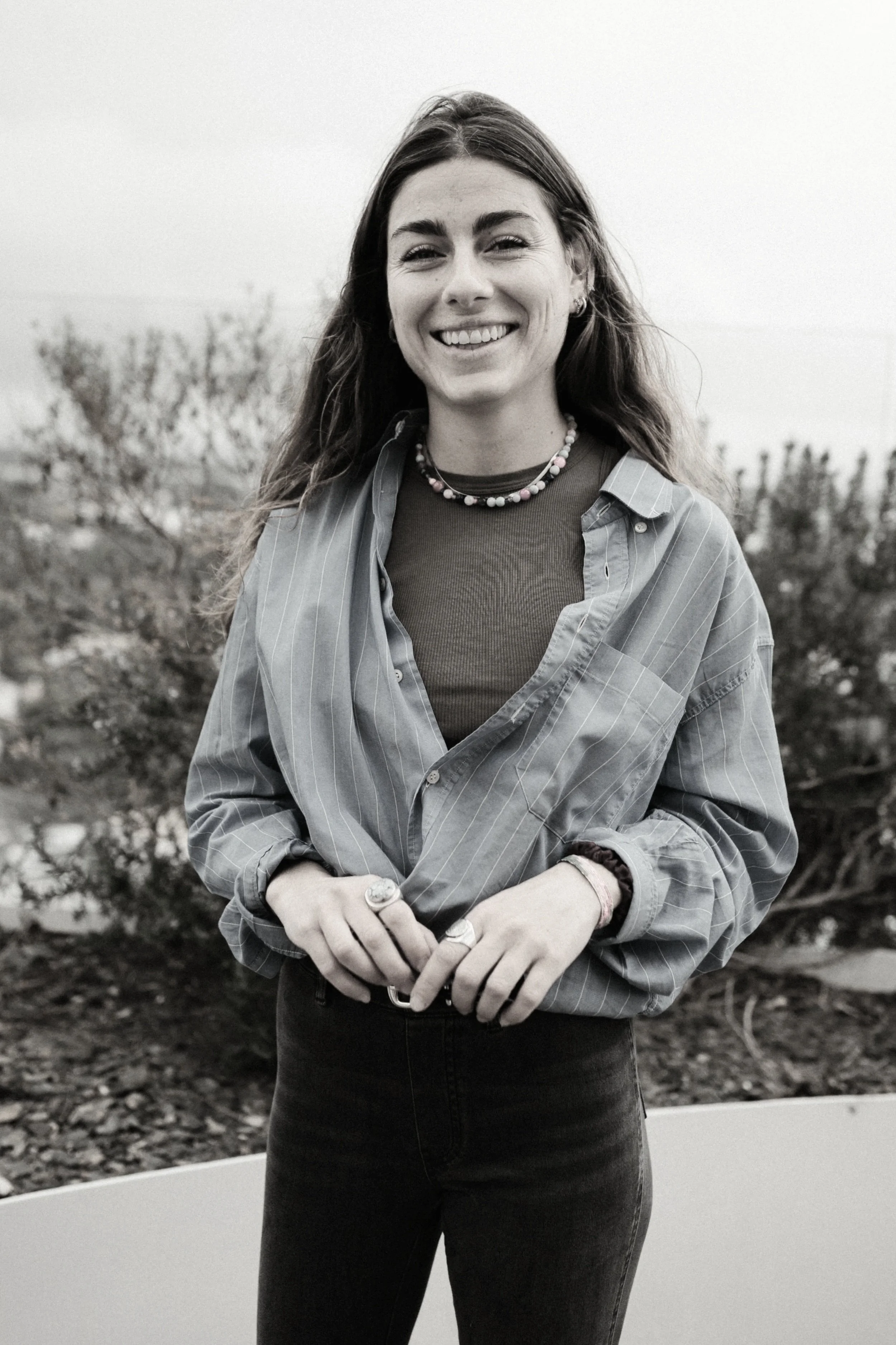 Une femme souriante avec les cheveux longs, portant un coli, un jean noir et des bijoux, debout à l'extérieur avec un paysage de buissons et un ciel clair.