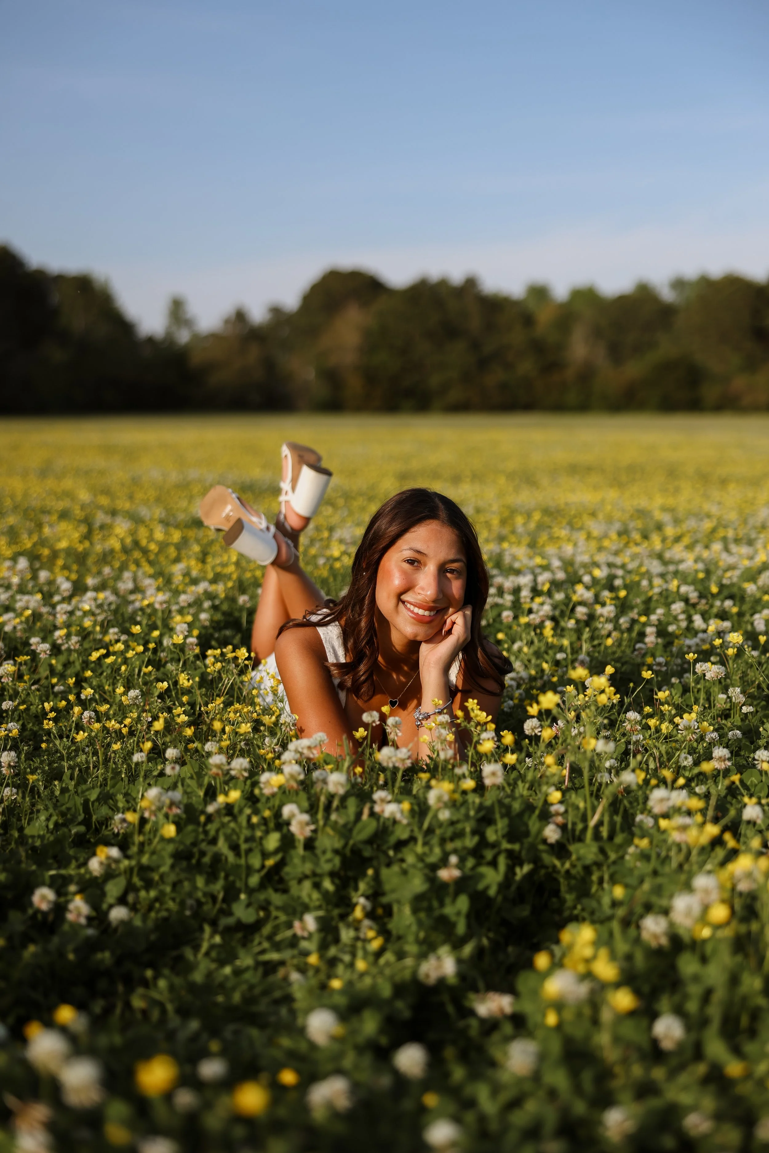 Young woman lying in a field of yellow and white flowers, smiling, with a backdrop of trees and a blue sky.