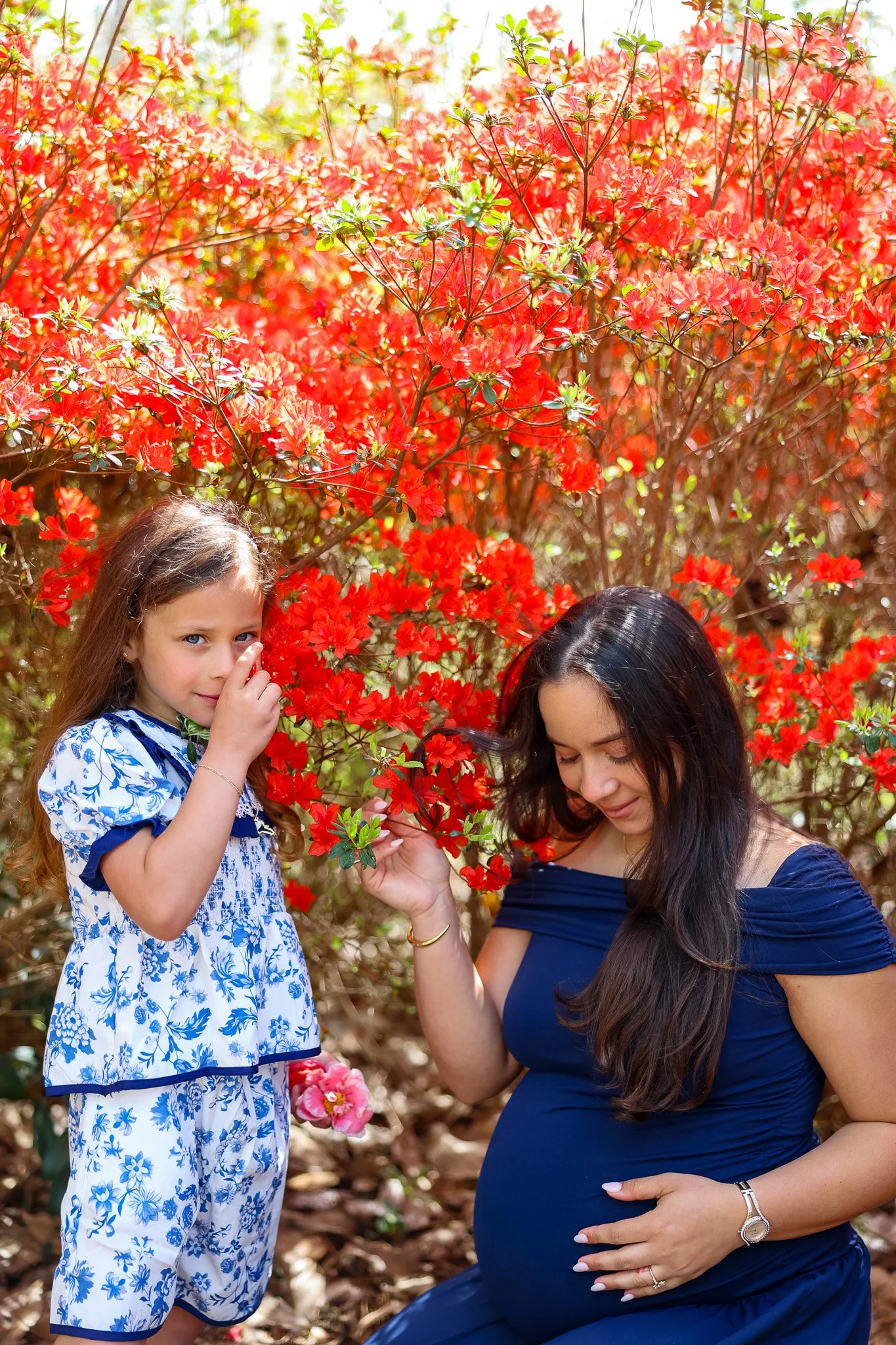 A woman with long dark hair in a navy blue dress is pregnant and sitting outdoors in front of a red flowering bush. A young girl with light brown hair and a blue and white floral dress is standing nearby, touching the bush and looking at the camera. The scene appears to be in a garden or park with sunlight filtering through the trees.