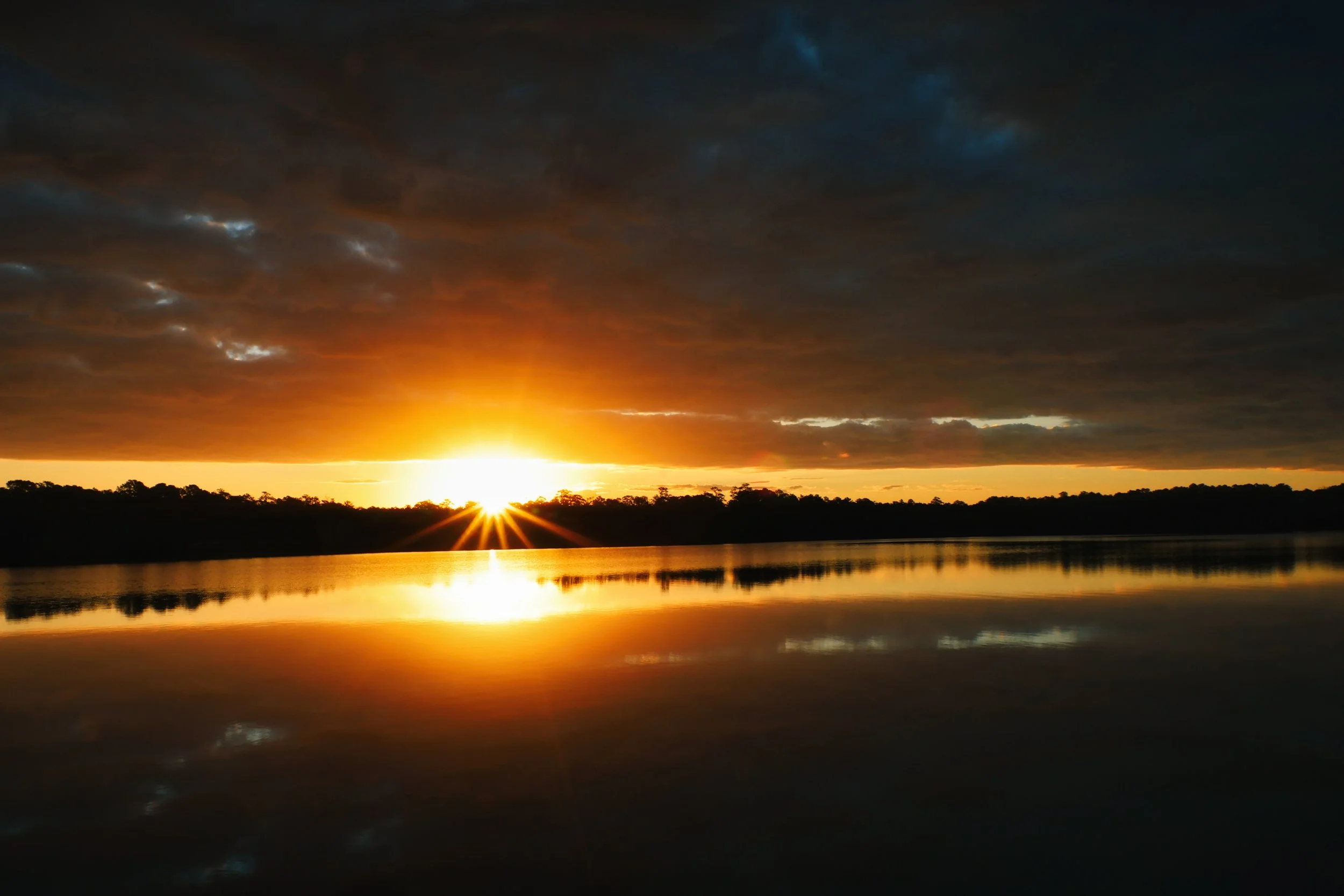 Sunset over a body of water with a dark cloudy sky, reflections on the water, and silhouettes of trees along the horizon.