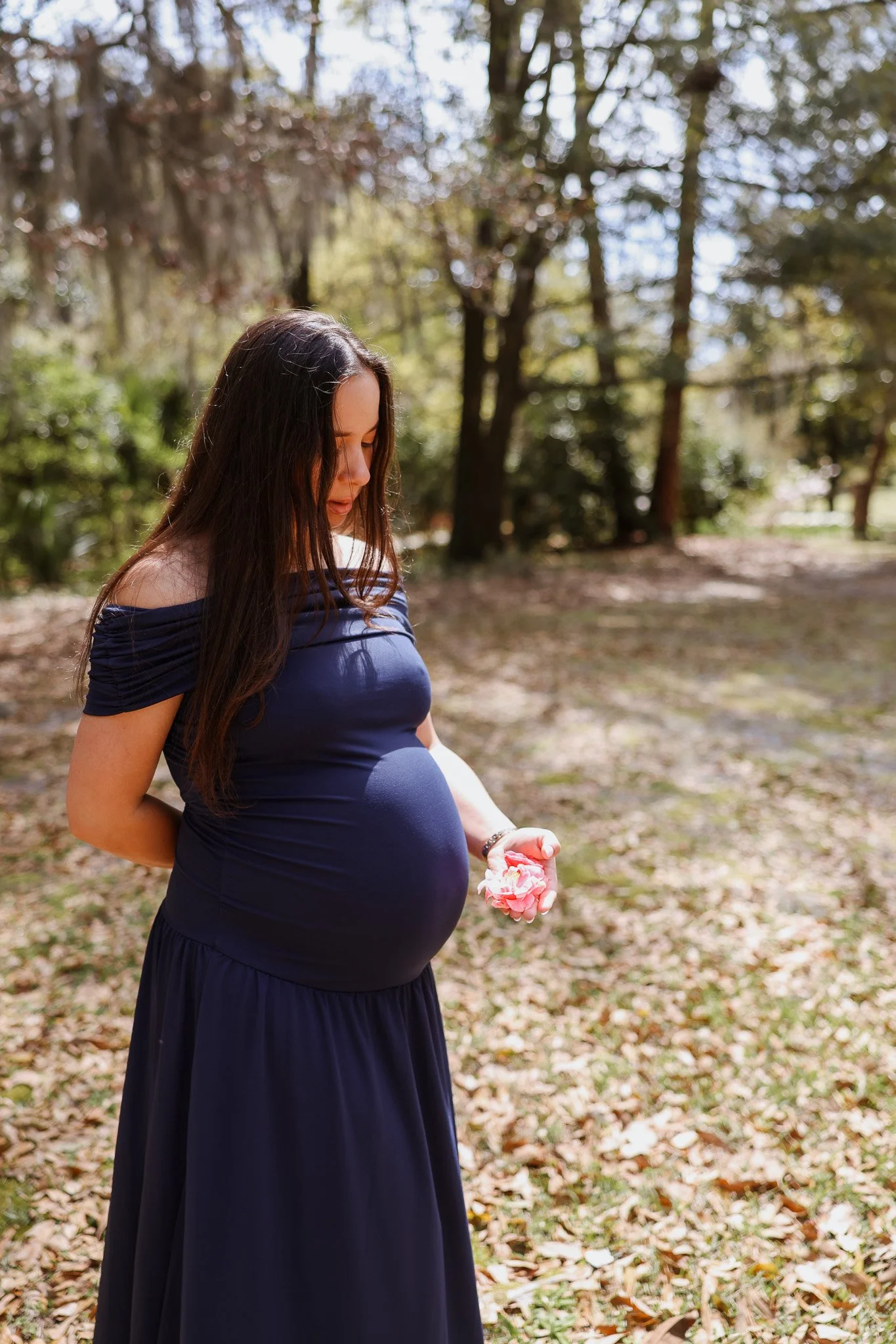 A pregnant woman outdoors holding pink flowers, wearing a dark blue off-shoulder dress in a wooded area with autumn leaves on the ground.