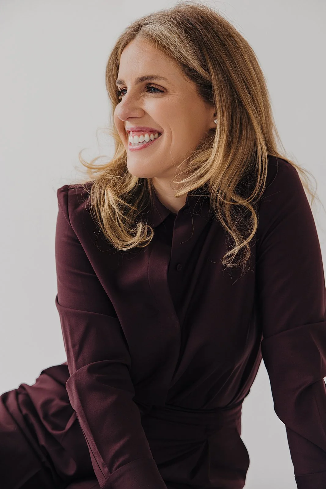 A woman with shoulder-length blonde hair smiling, dressed in a dark maroon blouse, sitting against a plain white background.