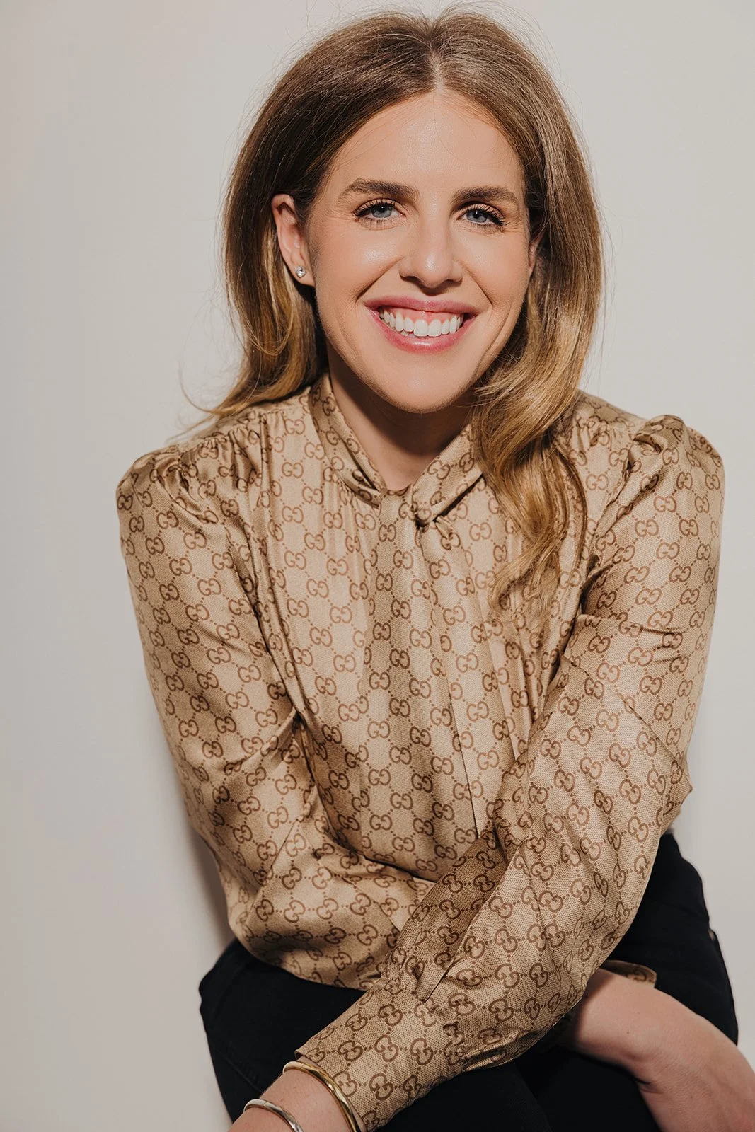 Woman with shoulder-length light brown hair, smiling, wearing a beige Gucci patterned blouse, sitting against a plain light background.
