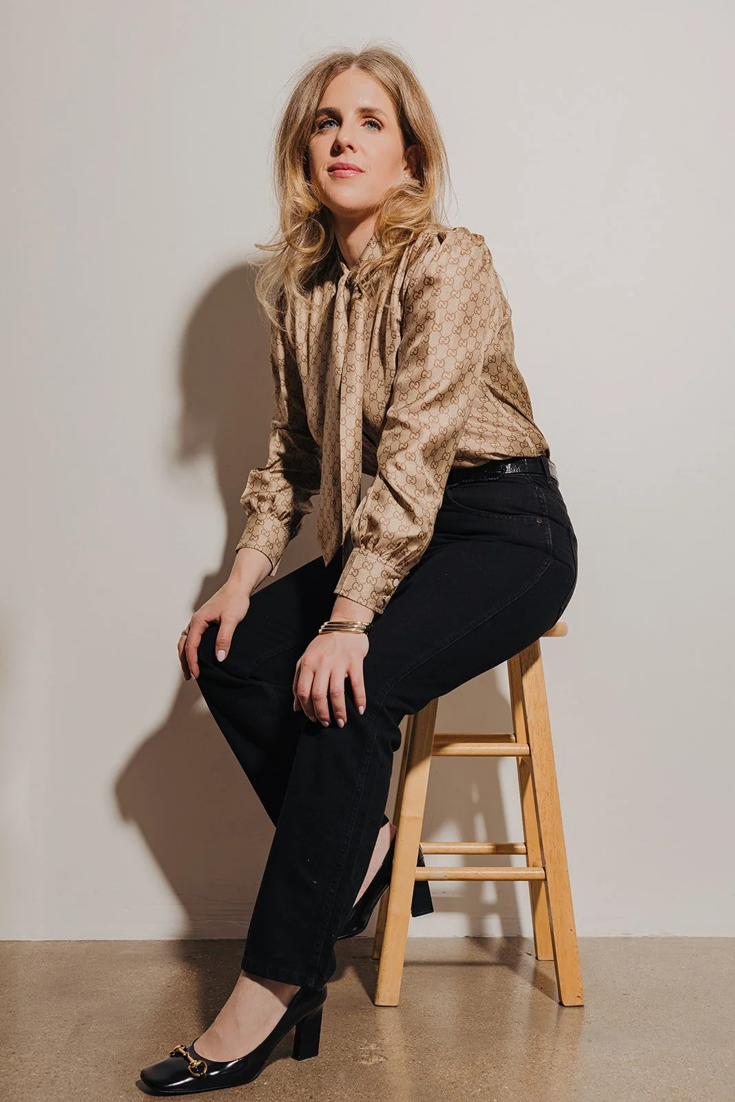A woman with blond hair sitting on a wooden stool, wearing a beige patterned blouse, black pants, and black heels, against a plain cream-colored background.