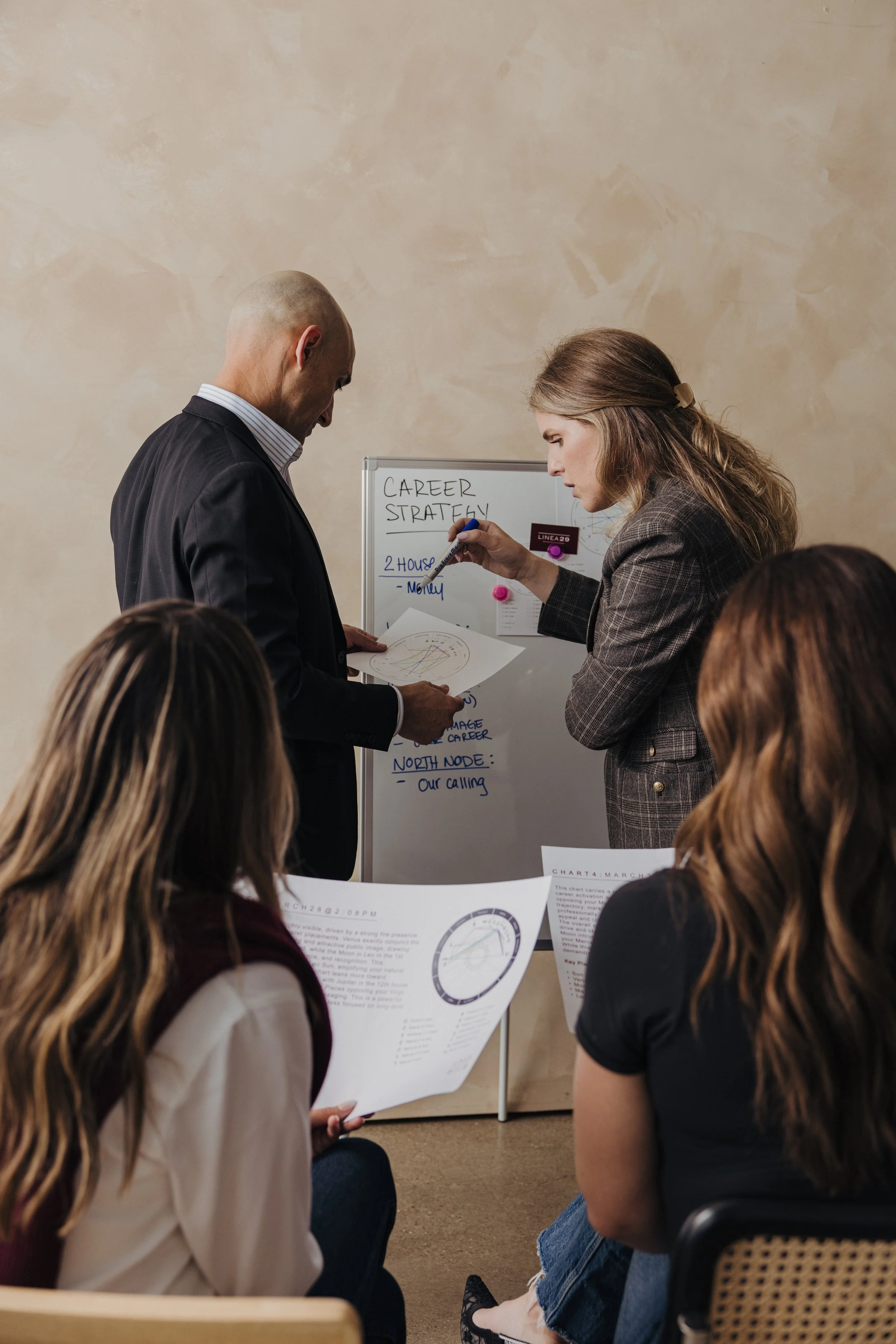 A group of people sitting in a room, listening to a woman in a plaid blazer explaining career strategy points written on a whiteboard. The whiteboard shows notes on career, money, and a north node.