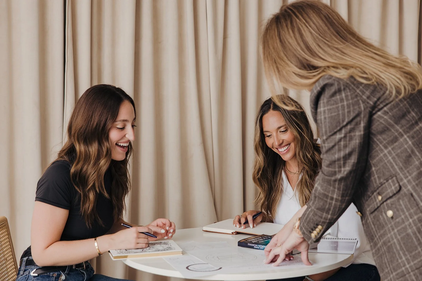 Three women sitting at a table engaged in a discussion, smiling, with notebooks and a book on finance or astronomy, one woman standing and leaning over the table.