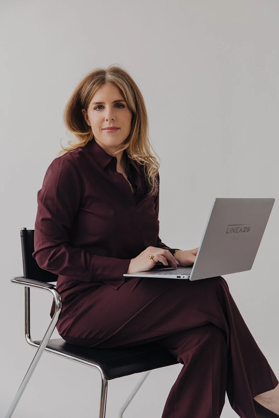 A woman with shoulder-length light brown hair and wearing a dark maroon shirt and pants, sitting on a black chair with metal legs, holding a silver Dell laptop on her lap, against a plain white background.