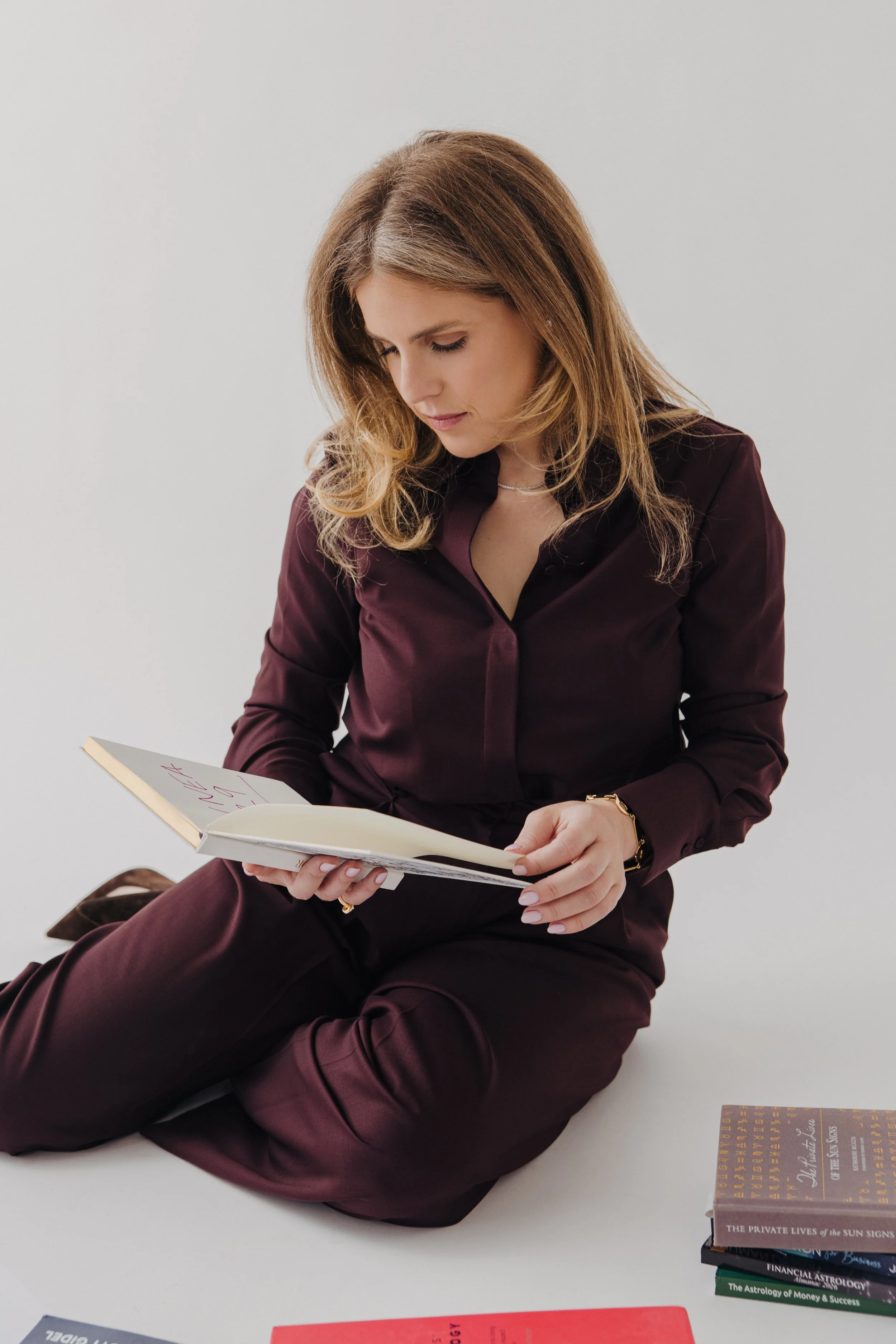 A woman with long, wavy hair wearing a dark burgundy suit reading a notebook or journal, sitting with crossed legs on a white surface. There are several stacked books on the table beside her.