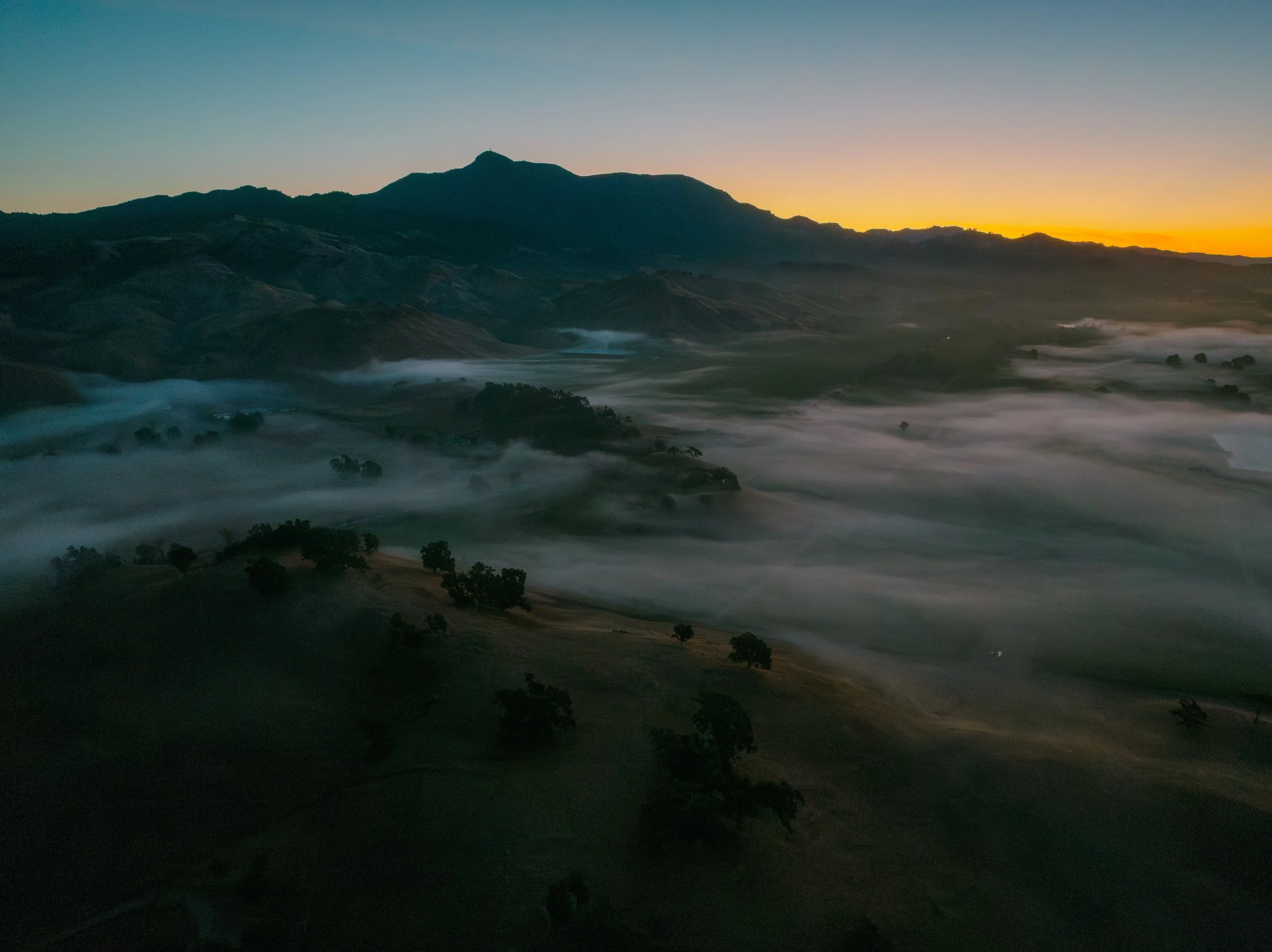 A mountain landscape at sunrise with a dark silhouette of mountain ranges, a clear sky, and mist or fog in the valleys.