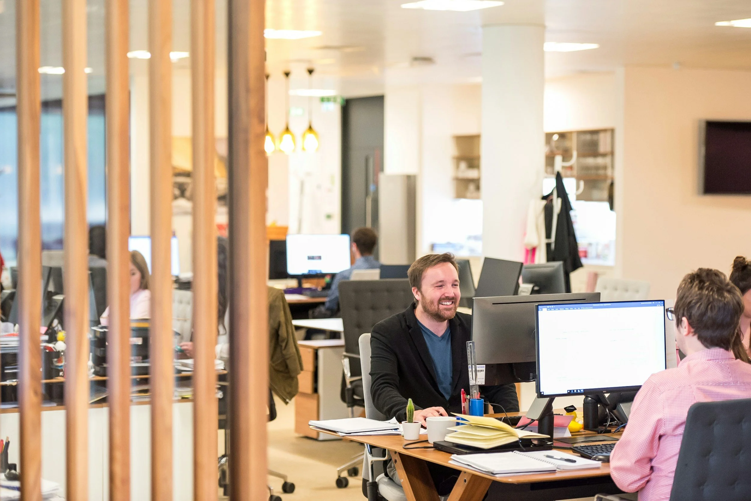 People working at desks in an open-plan office space with computers, papers, and office supplies, smiling and engaging with each other.