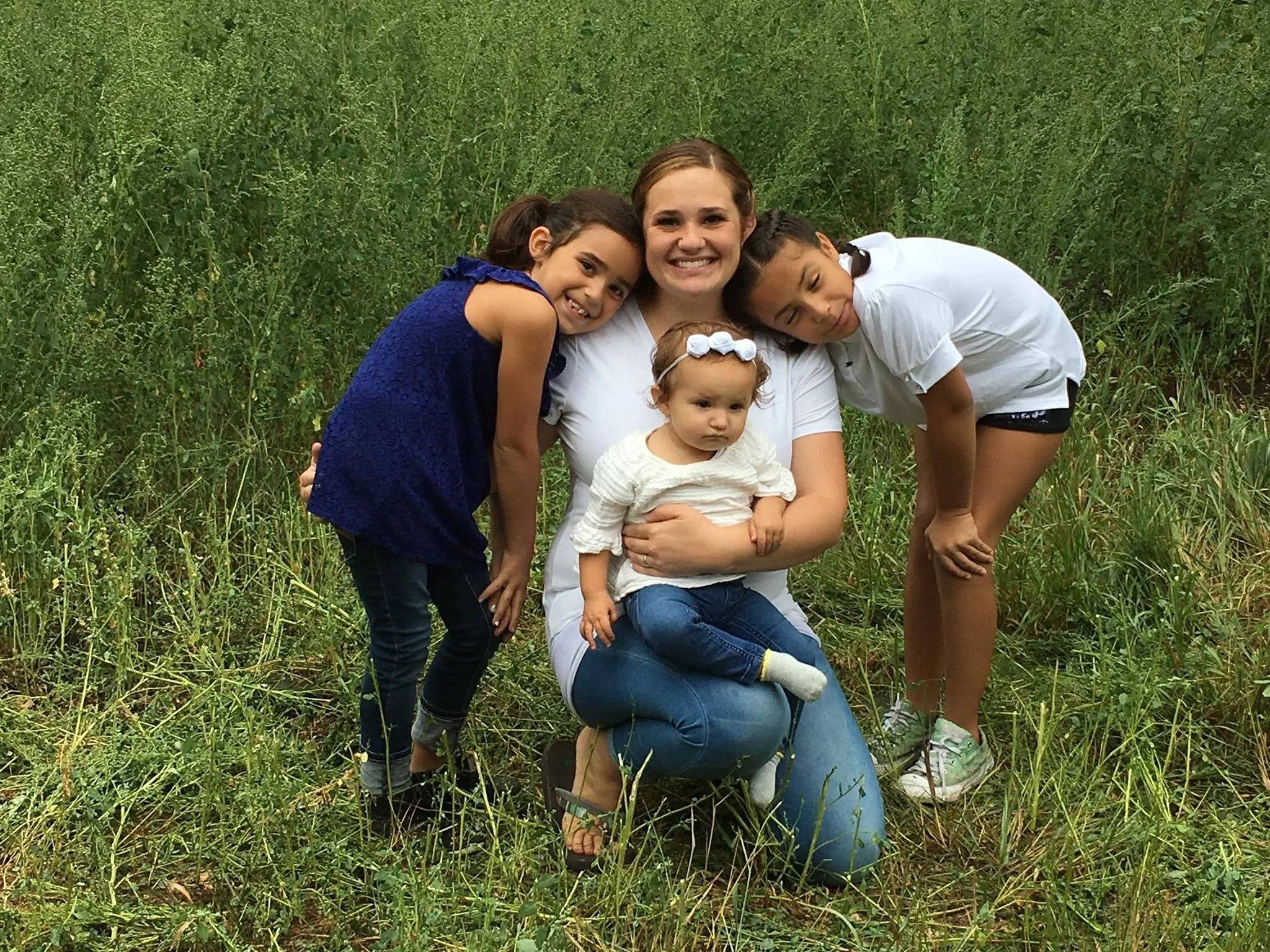 A group of five females, including a toddler, smiling and hugging each other in a grassy field surrounded by green foliage.