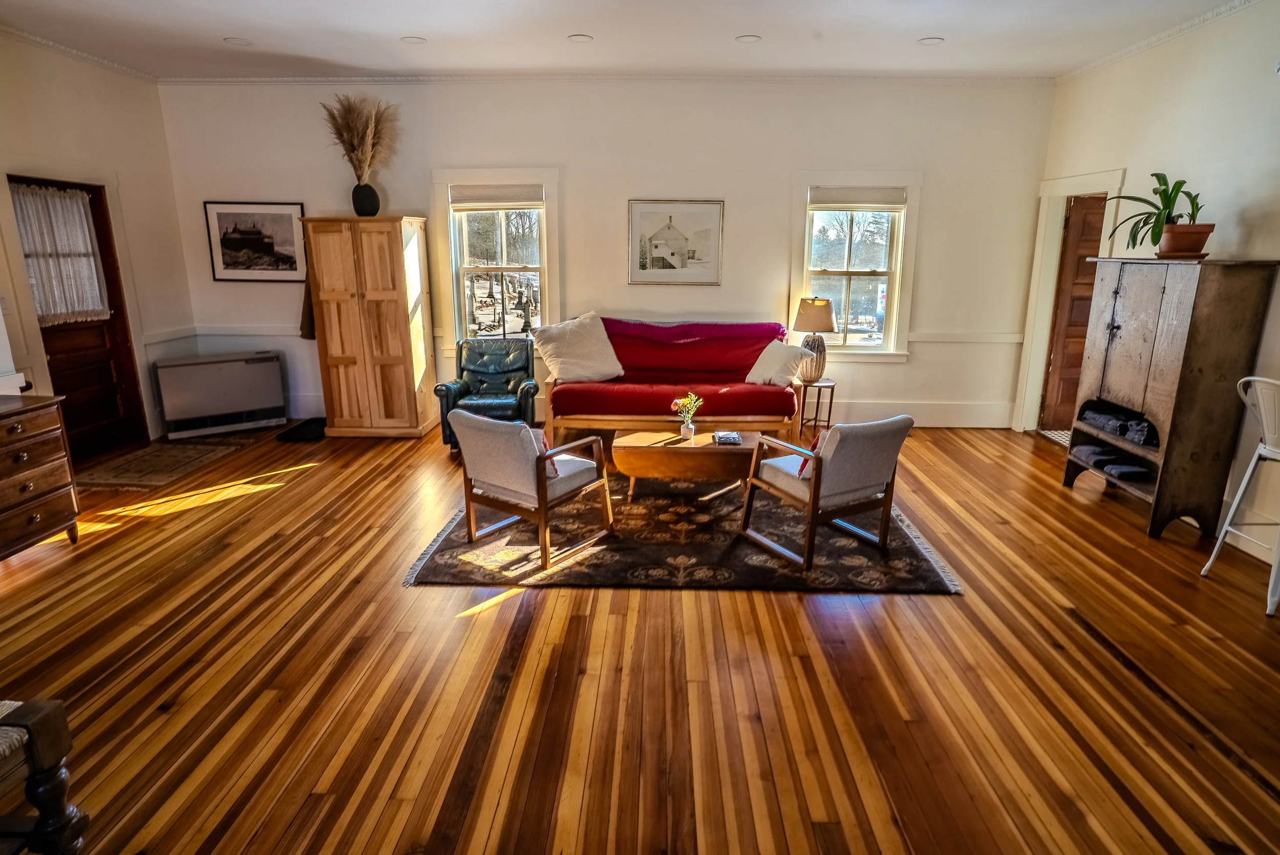 Living room with wooden floors, a red sofa, a black armchair, a small round coffee table, and windows with sunlight streaming in.