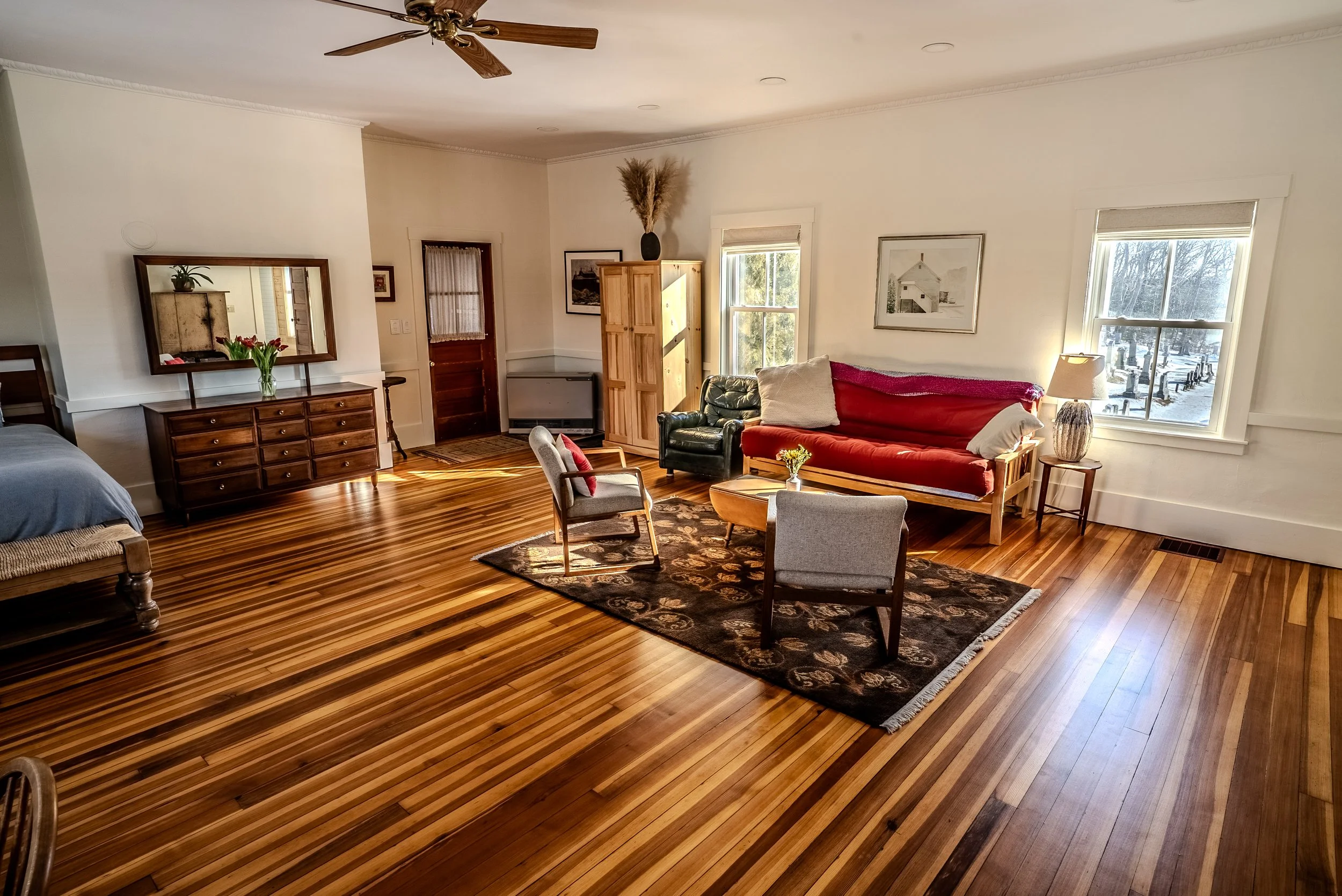 Living room with hardwood floors, a ceiling fan, a red couch, and multiple windows letting in natural light. There is a large mirror above a wooden dresser, framed artwork on the walls, and a black armchair with white and red pillows.