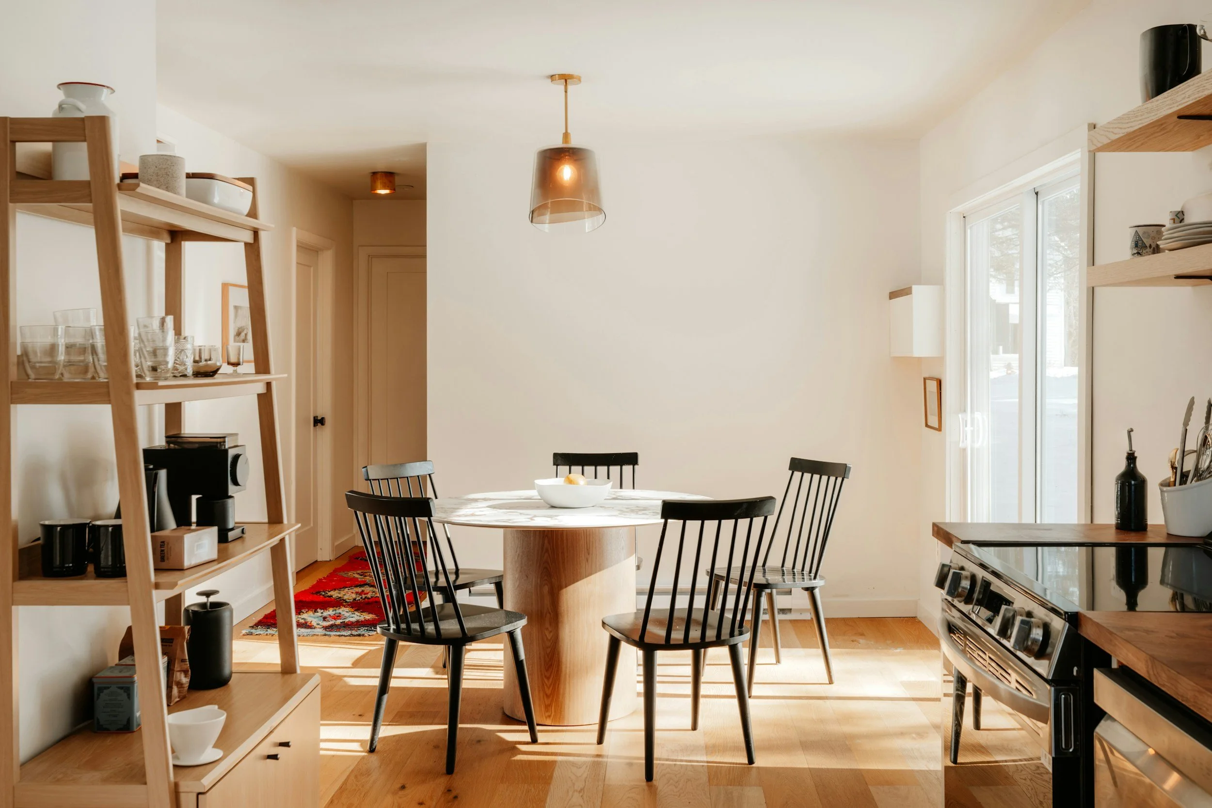 A cozy dining room with a round wooden table, black chairs, a white bowl with fruit, and a window with sunlight streaming in. There are shelves with dishes, a kitchen counter, and modern lighting fixtures.