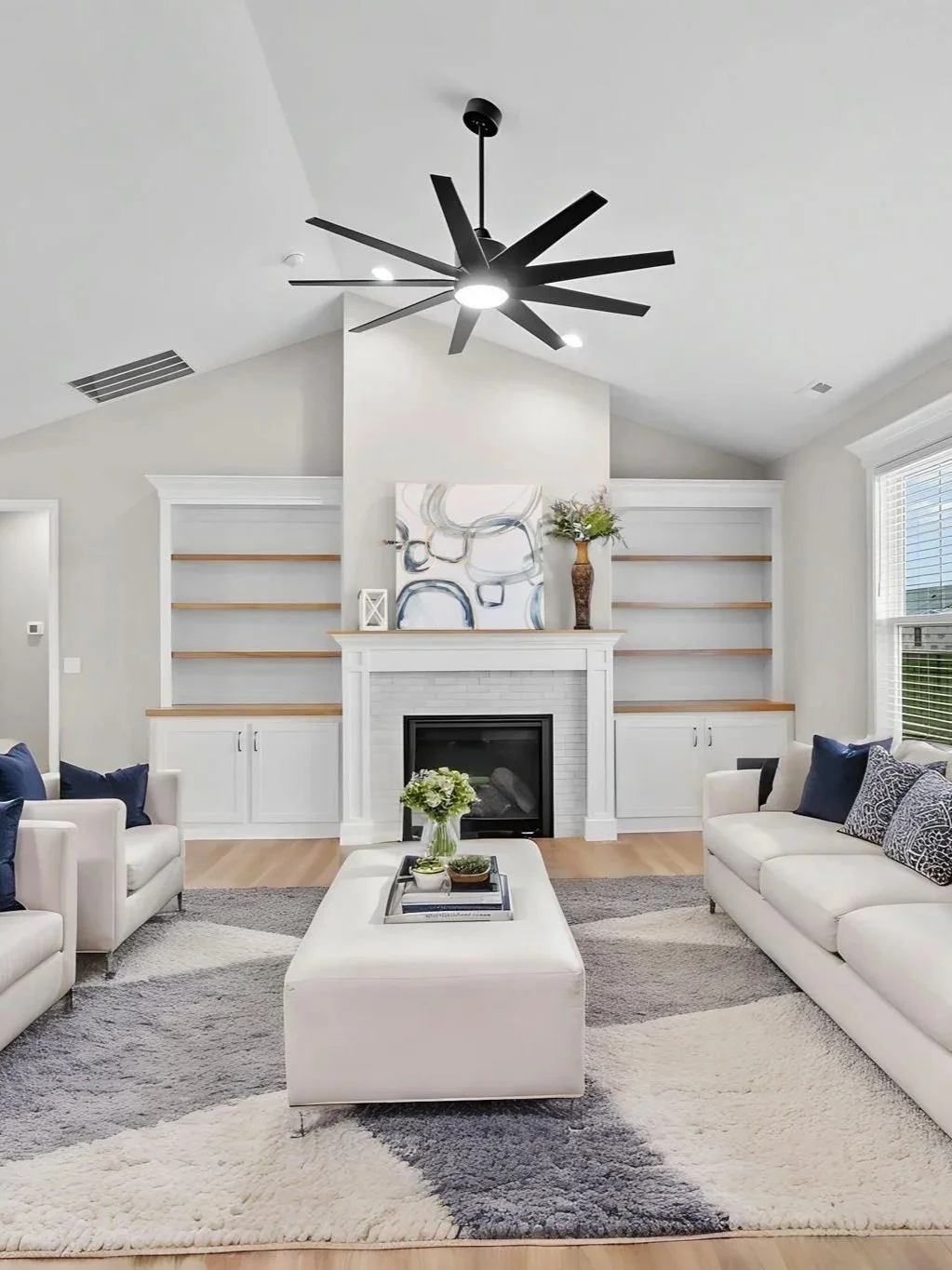 Living room with white sofas, a white rectangular ottoman, a fireplace, built-in shelves, a black ceiling fan, a large window, and modern decor.