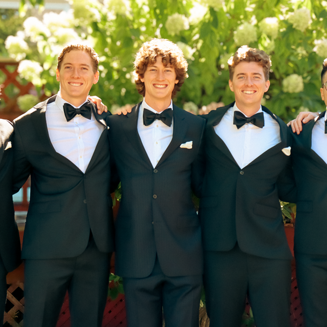 Four young men in tuxedos with bow ties, standing outdoors with green foliage and white flowers in the background, smiling with their arms around each other.