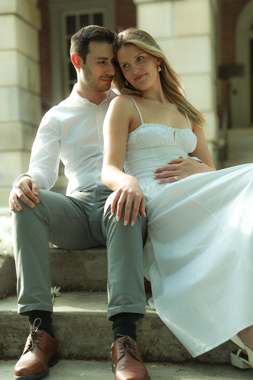 A man and a woman sitting close together on the steps outside a building, with their foreheads touching and the woman resting her hand on her stomach.