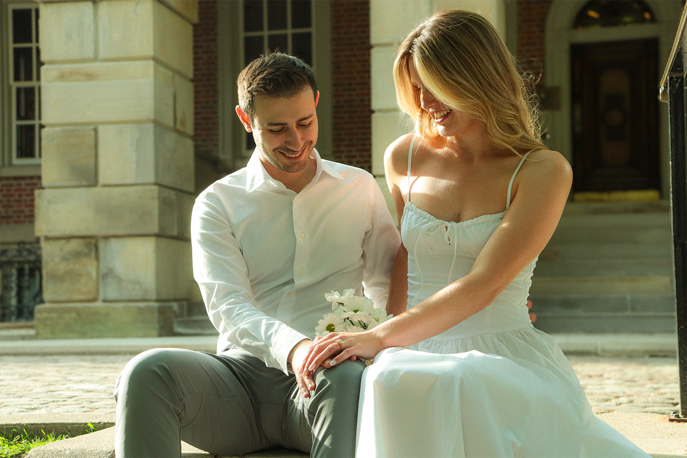 A man and woman sitting on the ground outside a building, smiling and holding hands. The woman is wearing a white dress, and the man is in a white shirt and gray pants. The scene appears to be romantic and joyful.