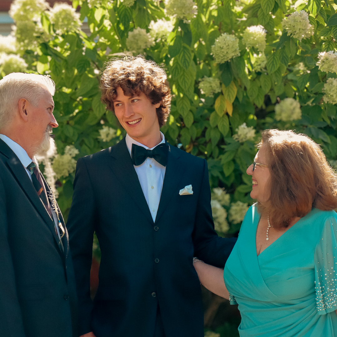 A young man in a tuxedo smiling while talking to an older man and woman outdoors near green foliage.