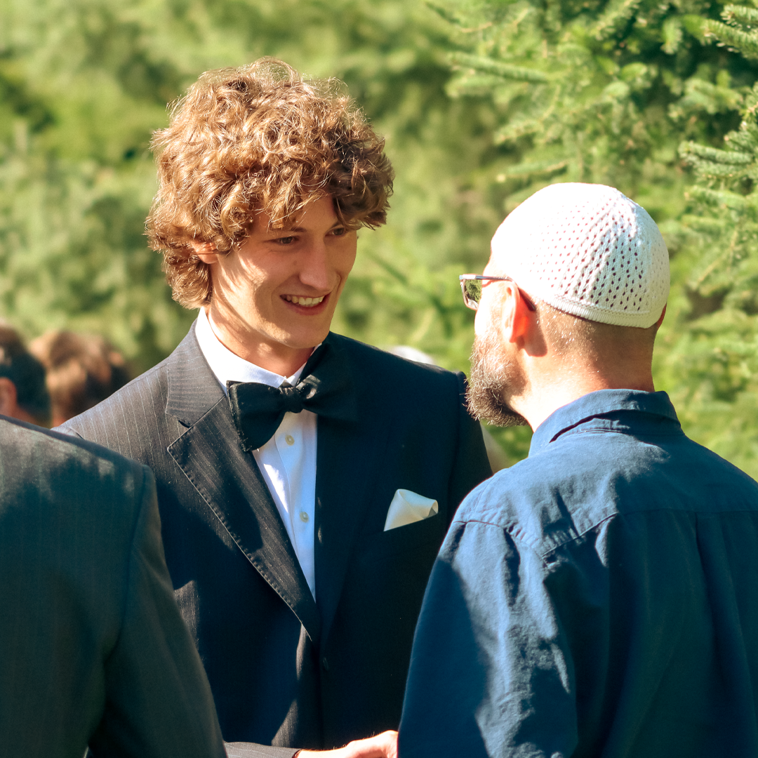 A young man with curly hair wearing a black tuxedo and bow tie, smiling and shaking hands with an older man in a blue shirt and white skullcap, outdoors with green trees in the background.