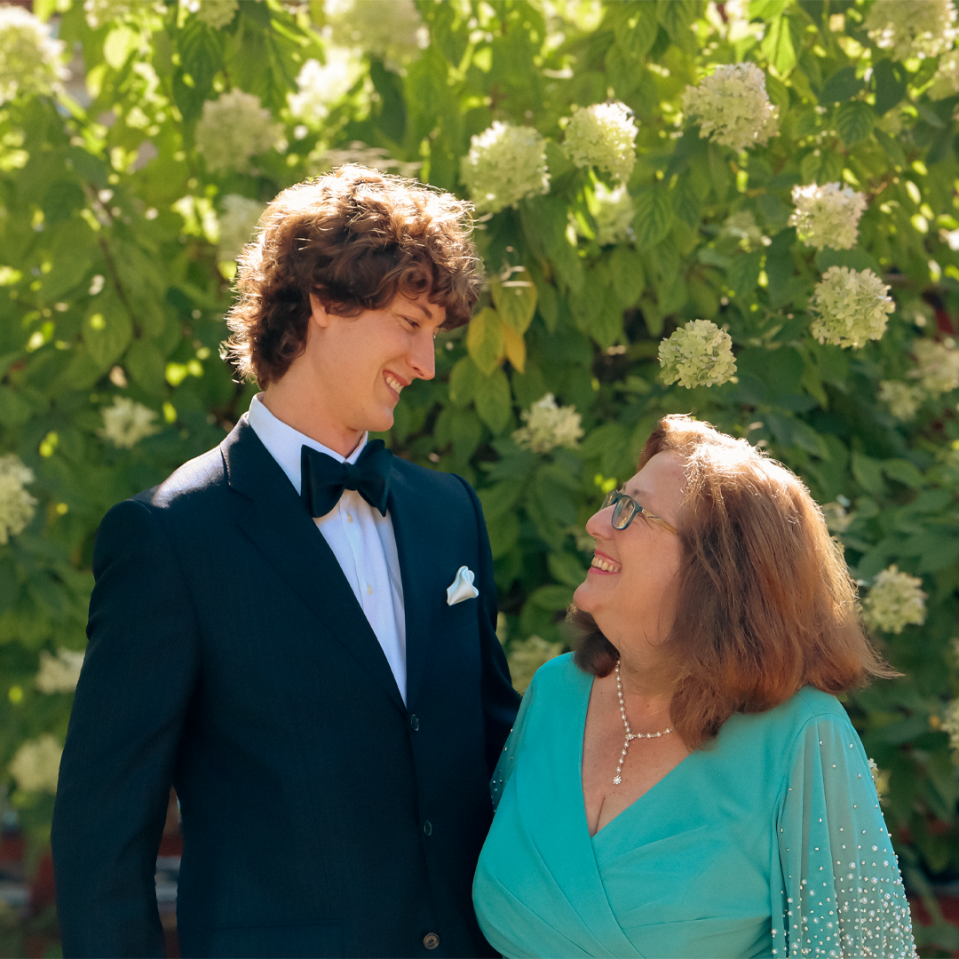 A young man in a tuxedo smiling at an older woman in a turquoise dress with pearl jewelry, outdoors with green foliage and white flowers in the background.