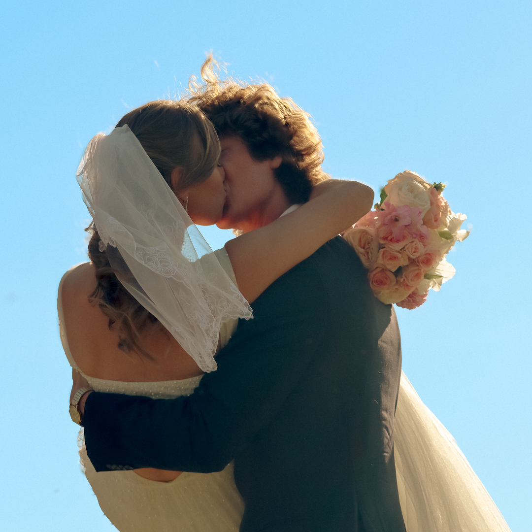 A bride and groom kissing outside during a wedding, with the bride holding a bouquet of pink and white flowers.