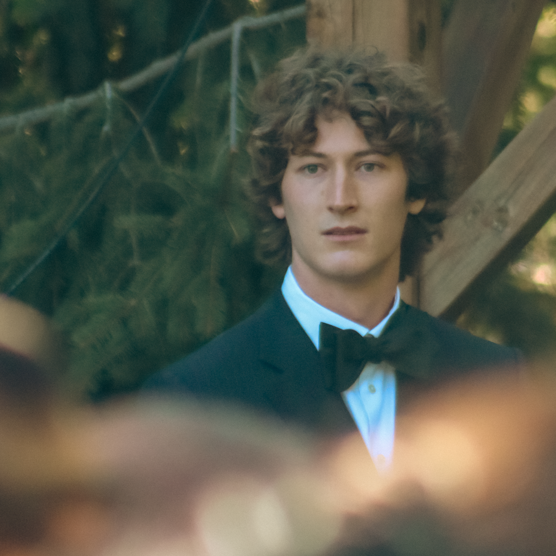A young man with curly hair in formal attire, wearing a tuxedo with a black bow tie, standing outdoors against a blurred background with trees and wooden structures.