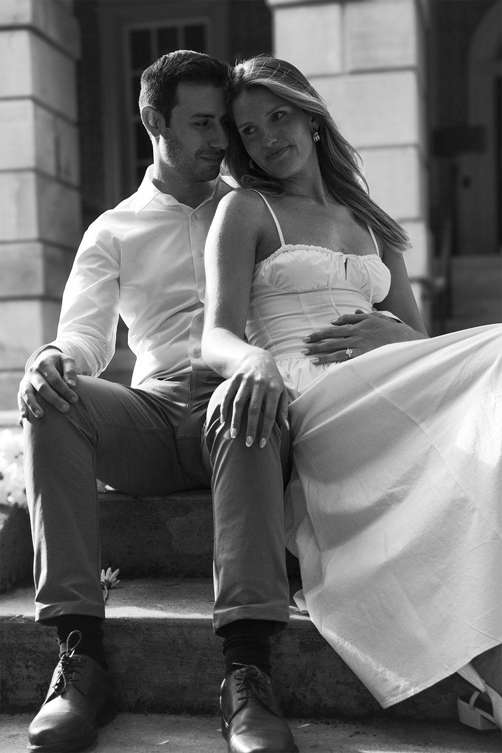 A black-and-white photo of a couple sitting on steps outside a building. The man is wearing a white shirt and light-colored pants, and the woman is wearing a light-colored dress. They are close together, with the man gently holding the woman's waist 