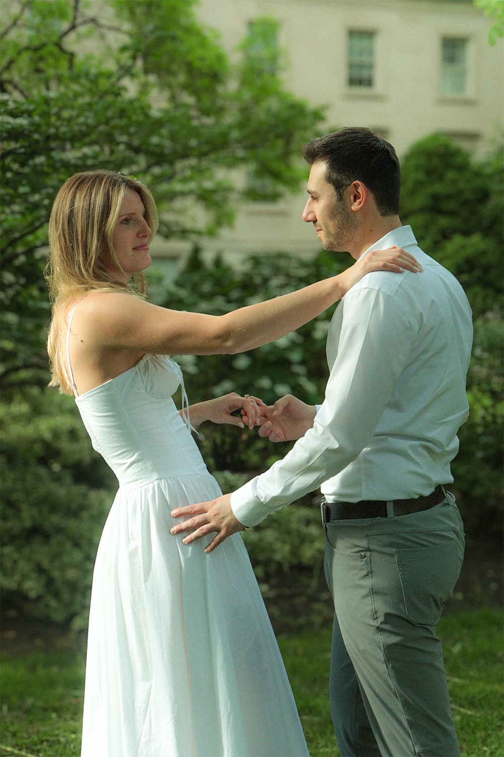A couple dressed in white, holding hands and looking into each other's eyes outdoors on a green, leafy background.
